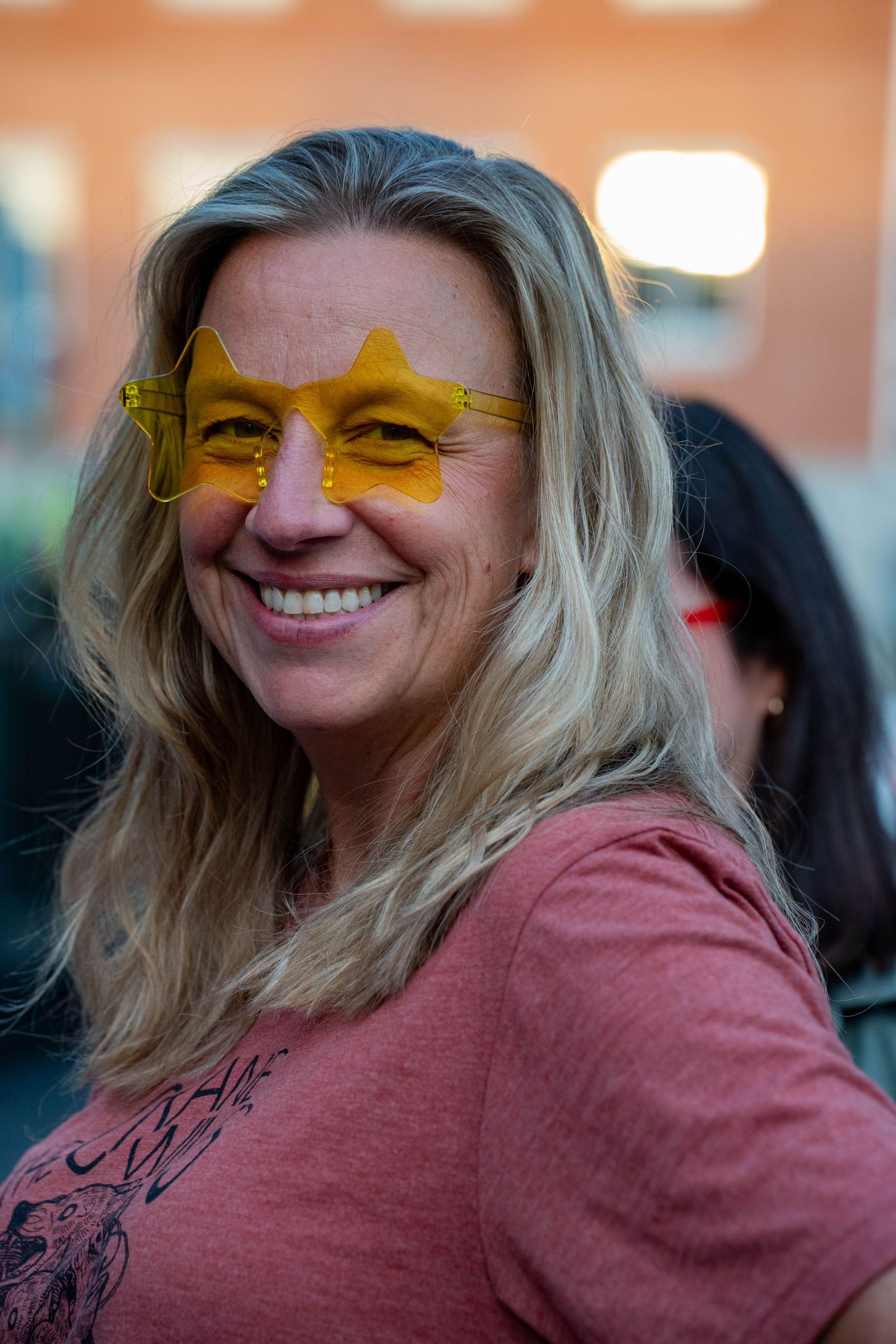 Smiling blonde woman wearing fun yellow star-shaped sunglasses and a pink t-shirt at an outdoor event.