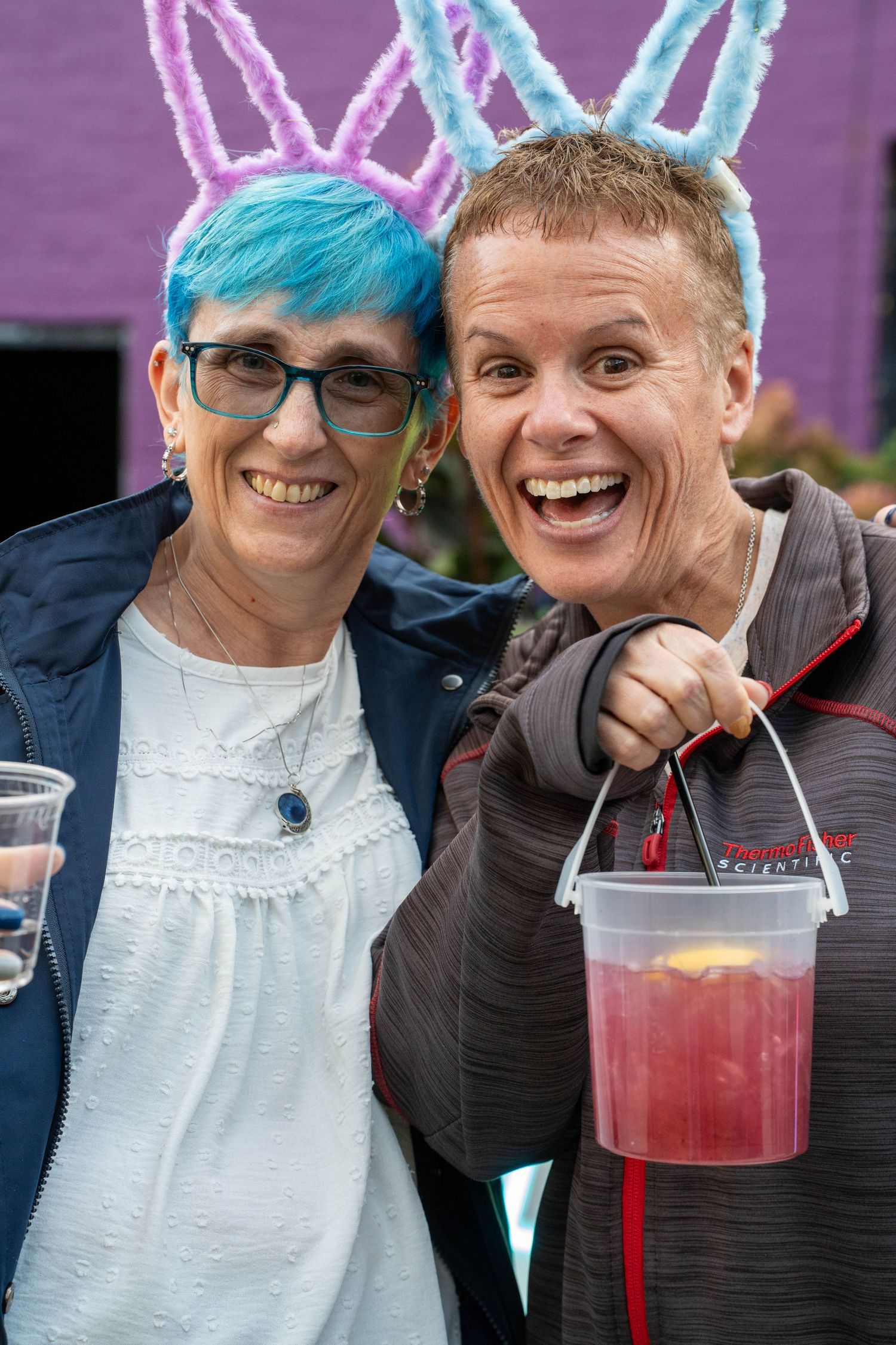Two people wearing bunny ear headbands smile at camera, one holding a pink drink at an outdoor Easter event.