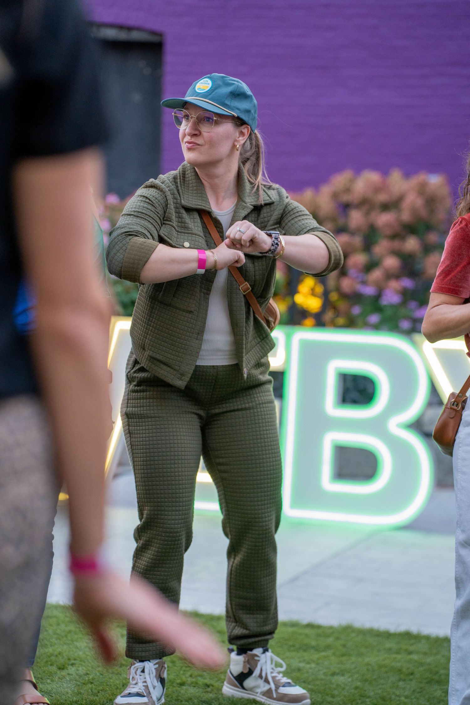Woman in olive green outfit and teal cap checks her watch at an outdoor event with purple wall backdrop.