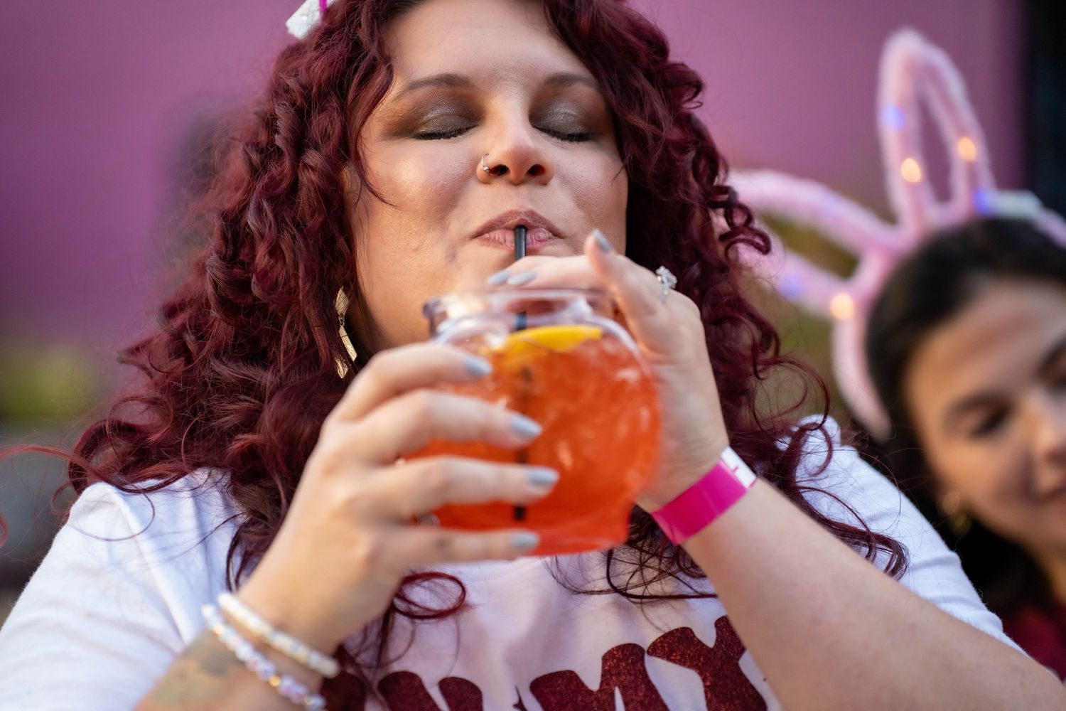 Woman with curly red hair sipping an orange cocktail through a straw at an outdoor event.