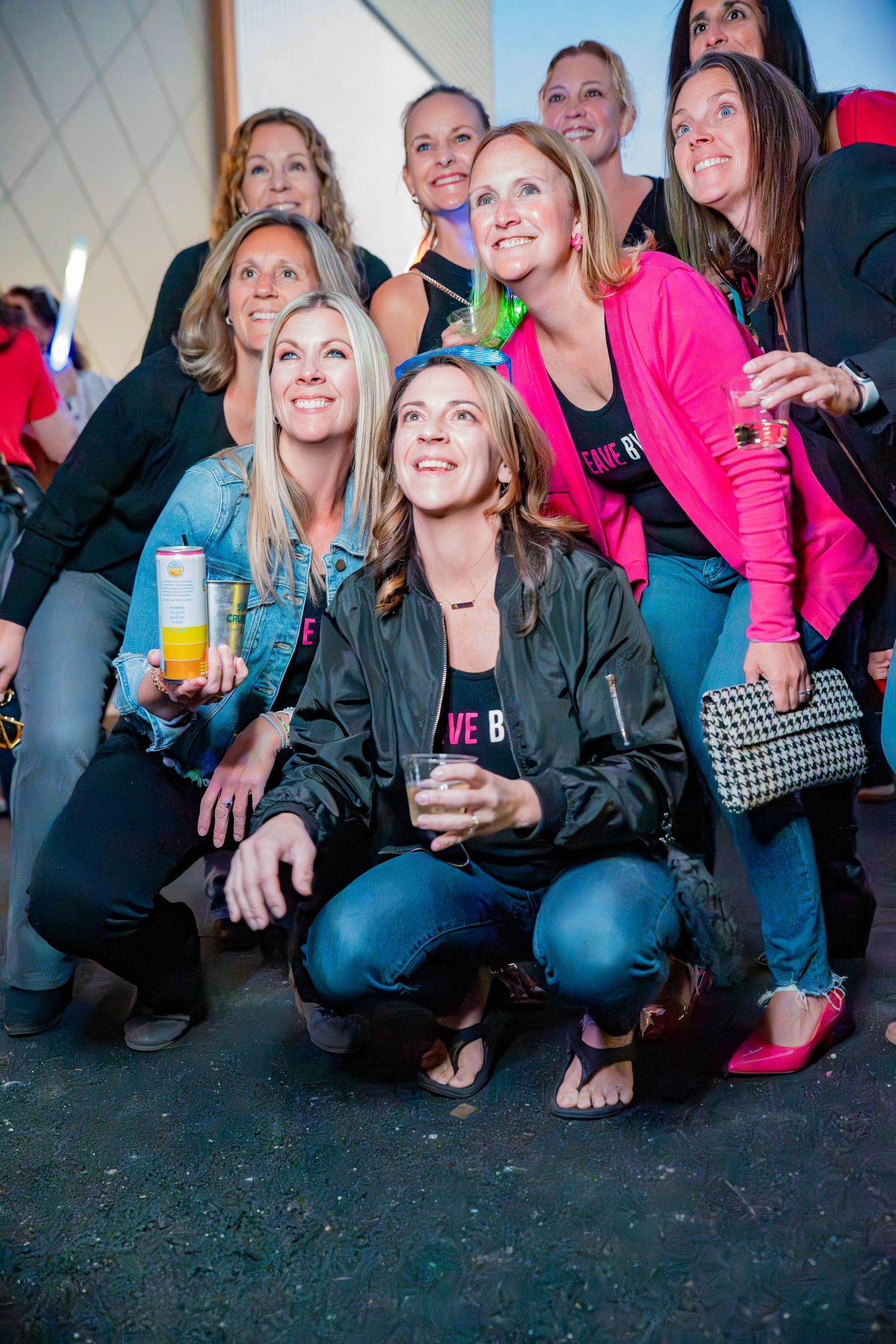 Group of women smiling and posing together at a lively evening event, some holding drinks, dressed in casual attire.