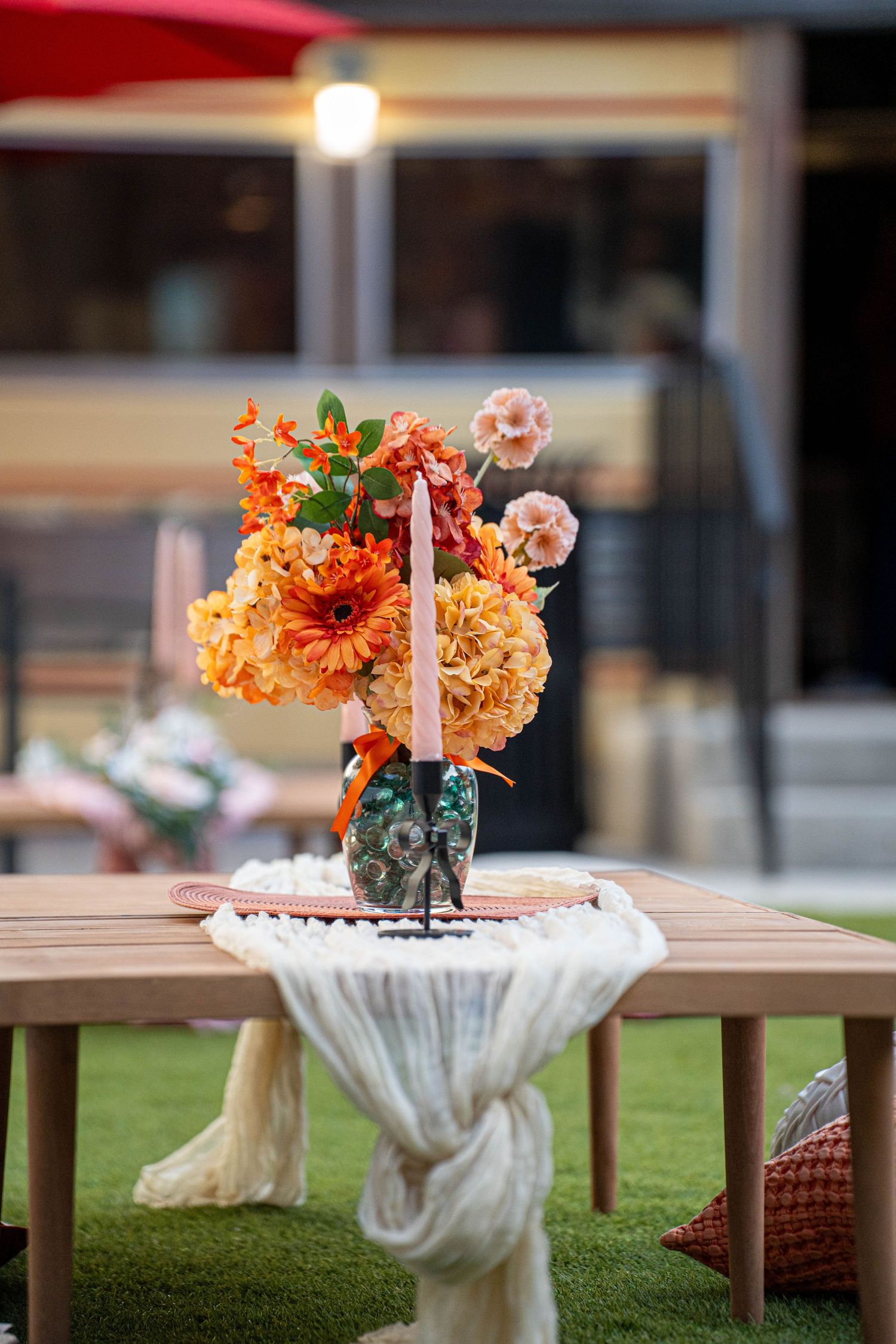 Elegant floral centerpiece with orange and peach blooms on wooden table with white fabric runner outdoors.