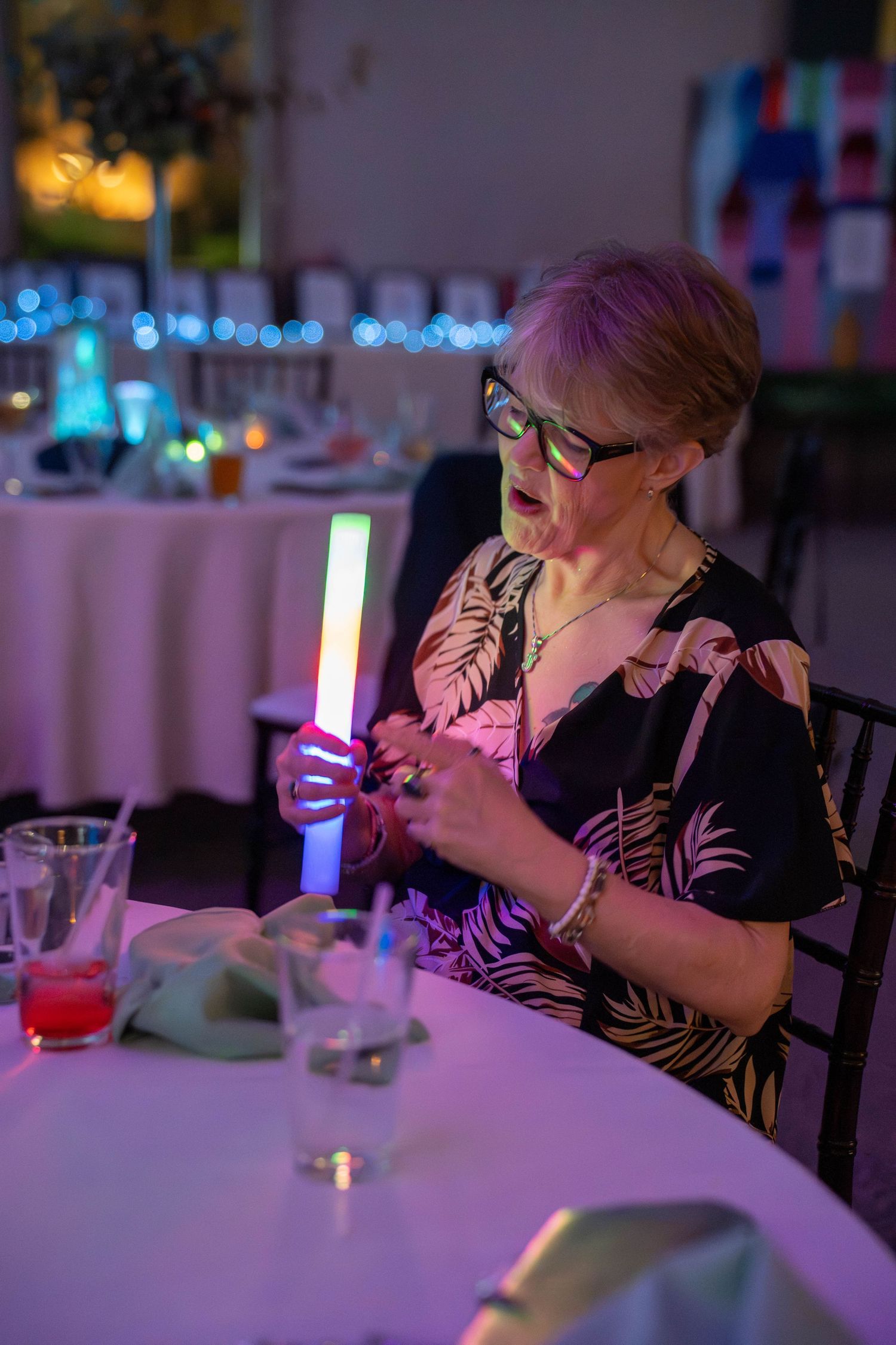 Elderly woman with pink hair and green glasses holds a glowing LED stick at a festive event dinner table.