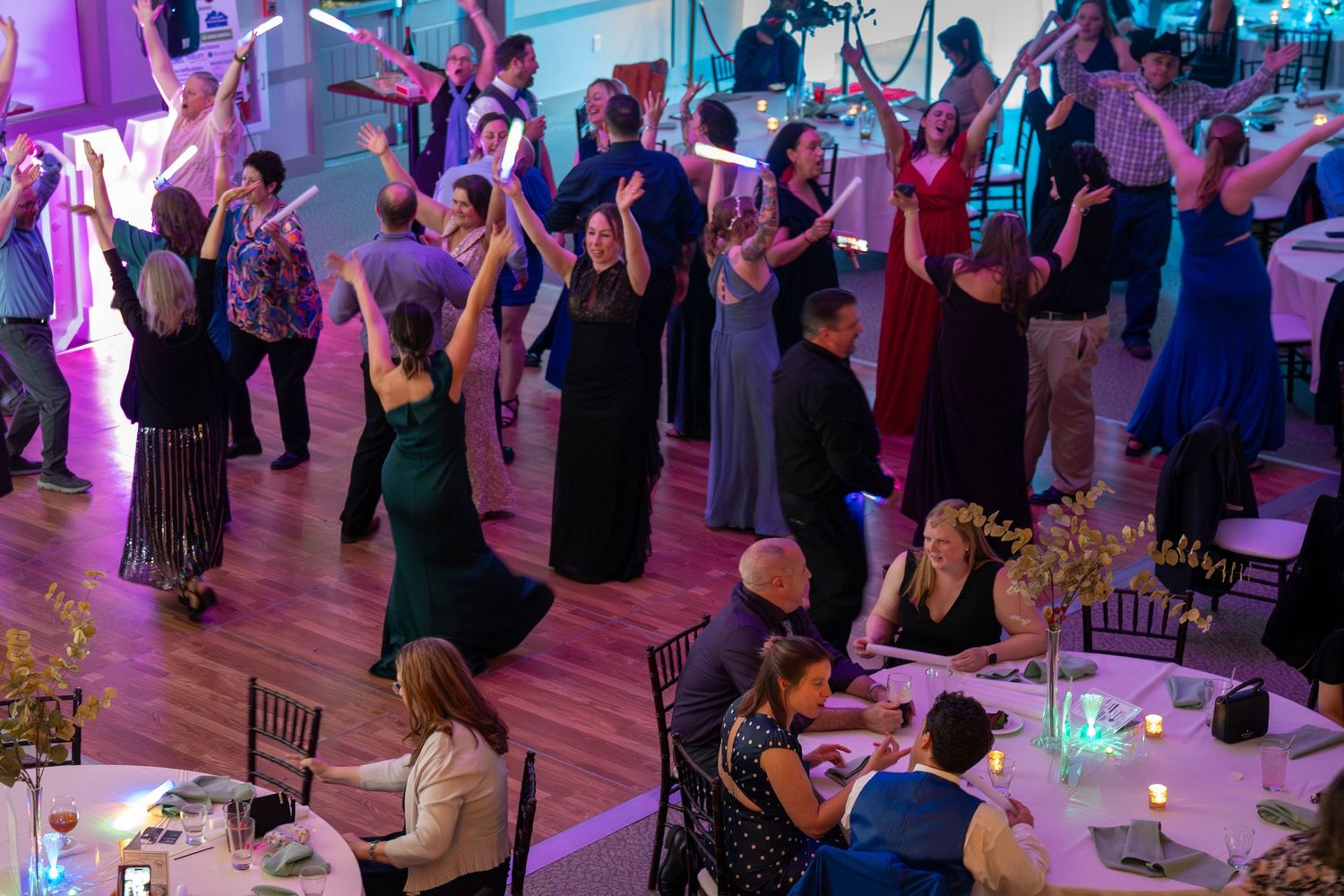 Guests dancing with arms raised at an elegant wedding reception with purple lighting and decorated dining tables.