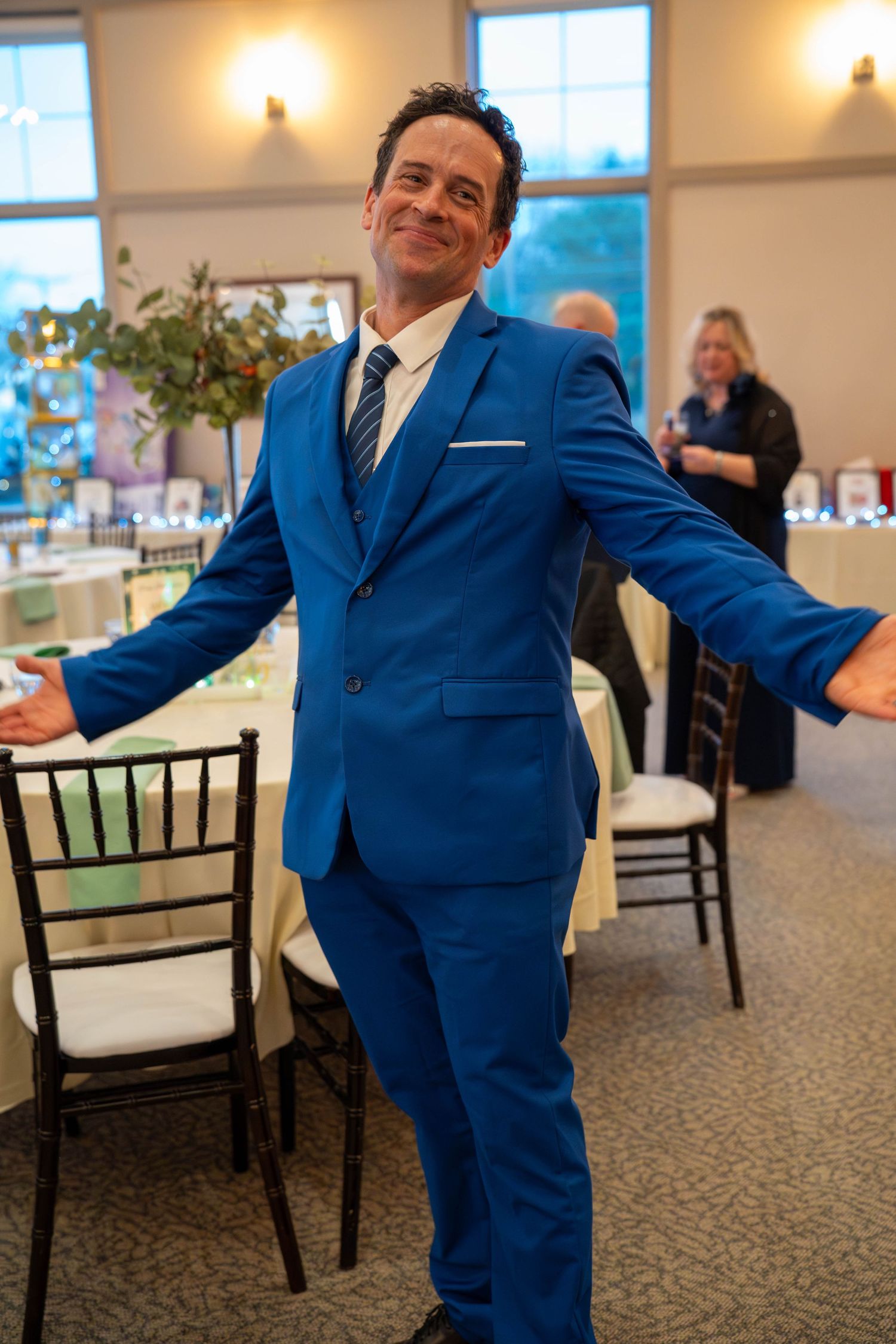 Man in royal blue three-piece suit with arms outstretched smiling at indoor wedding reception venue.