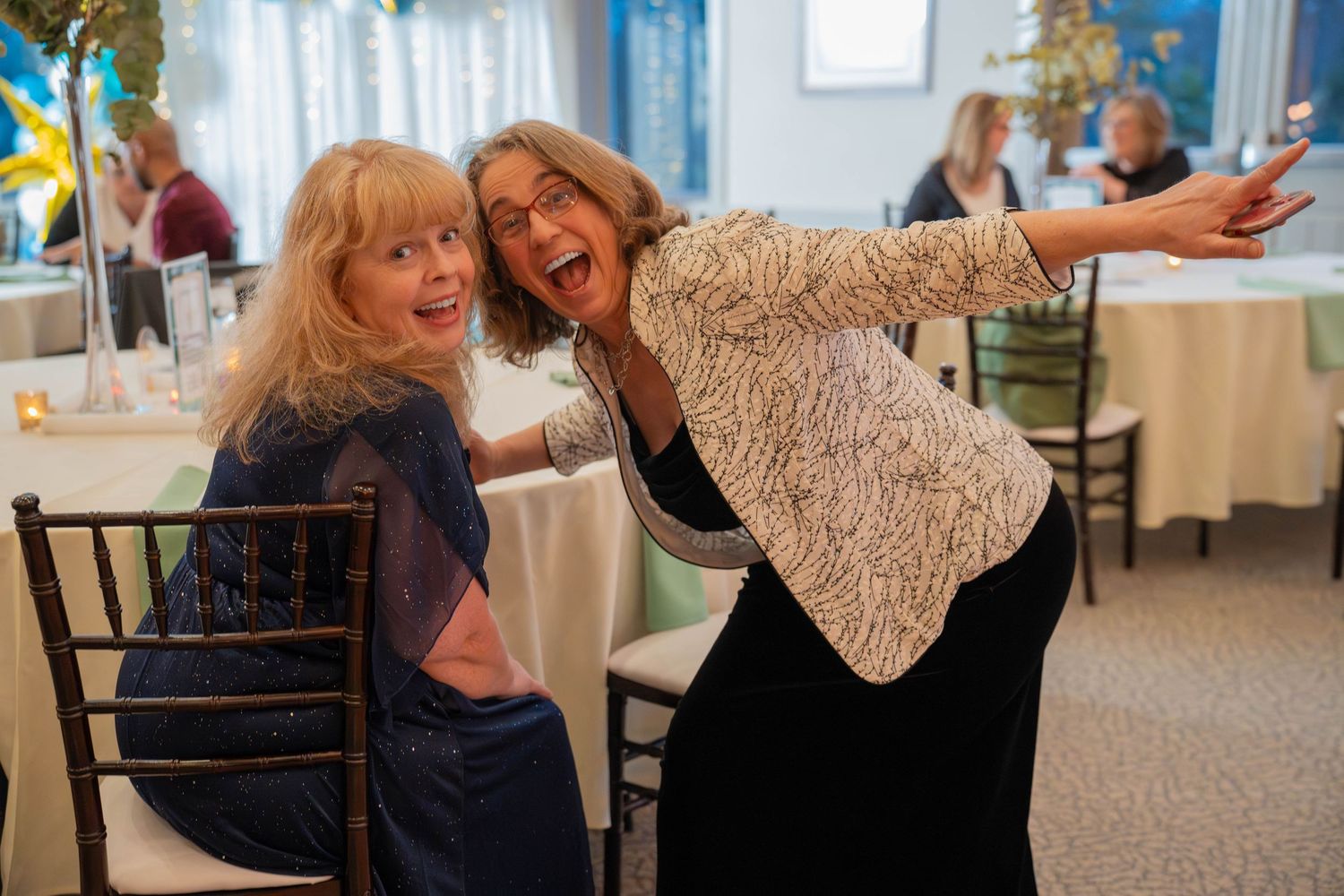 Two women laughing and posing joyfully at an elegant indoor event with white tablecloths and decorations.