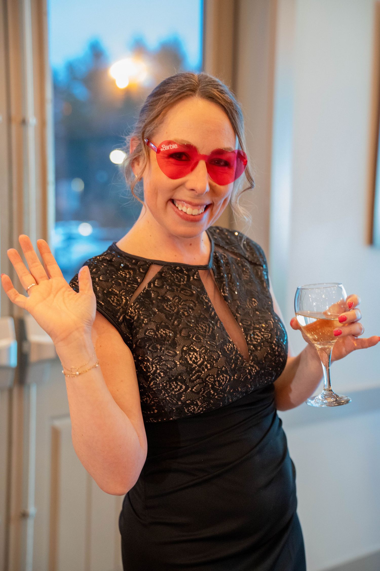 Smiling woman in black lace dress and red heart sunglasses waving while holding a glass of rosé wine.