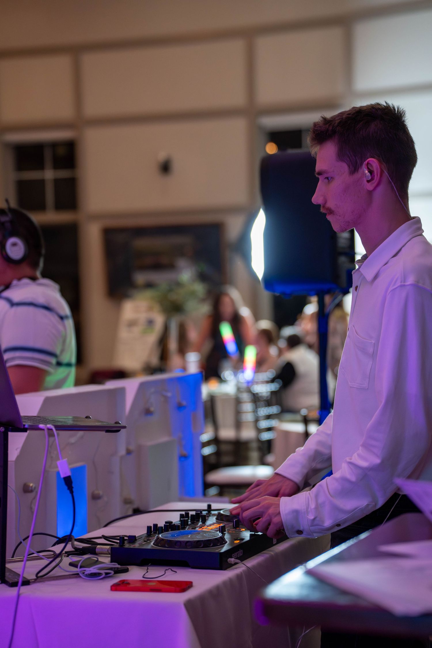 Young male DJ in white shirt operates mixing equipment at an elegant indoor event with colorful lighting.