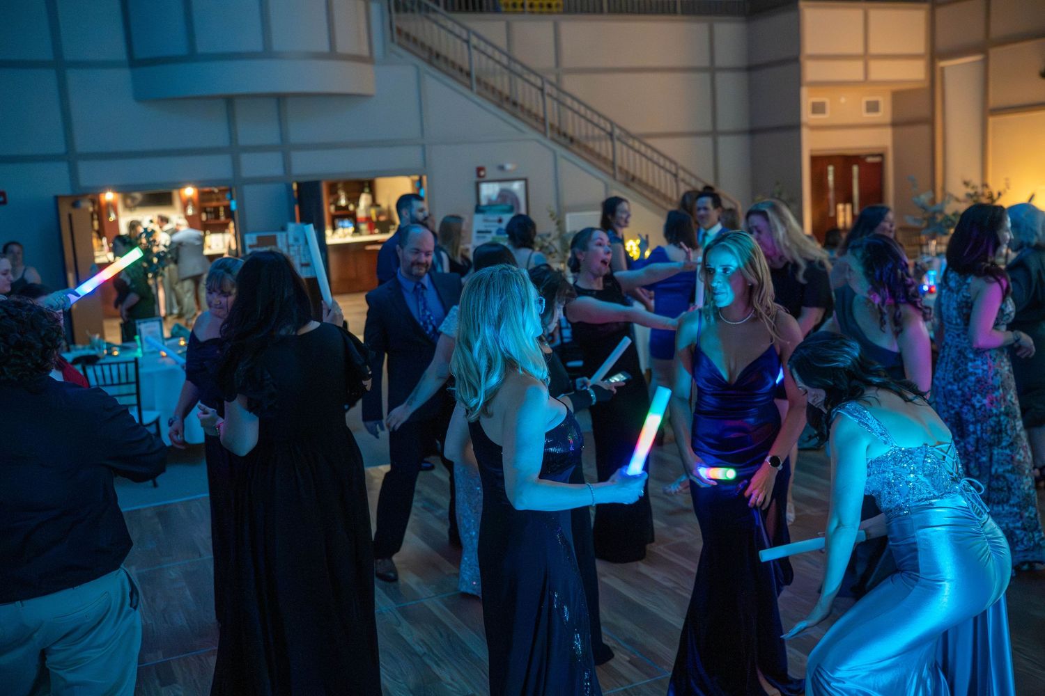 Guests dance at an elegant evening gala, holding glowing green light sticks in a grand venue.