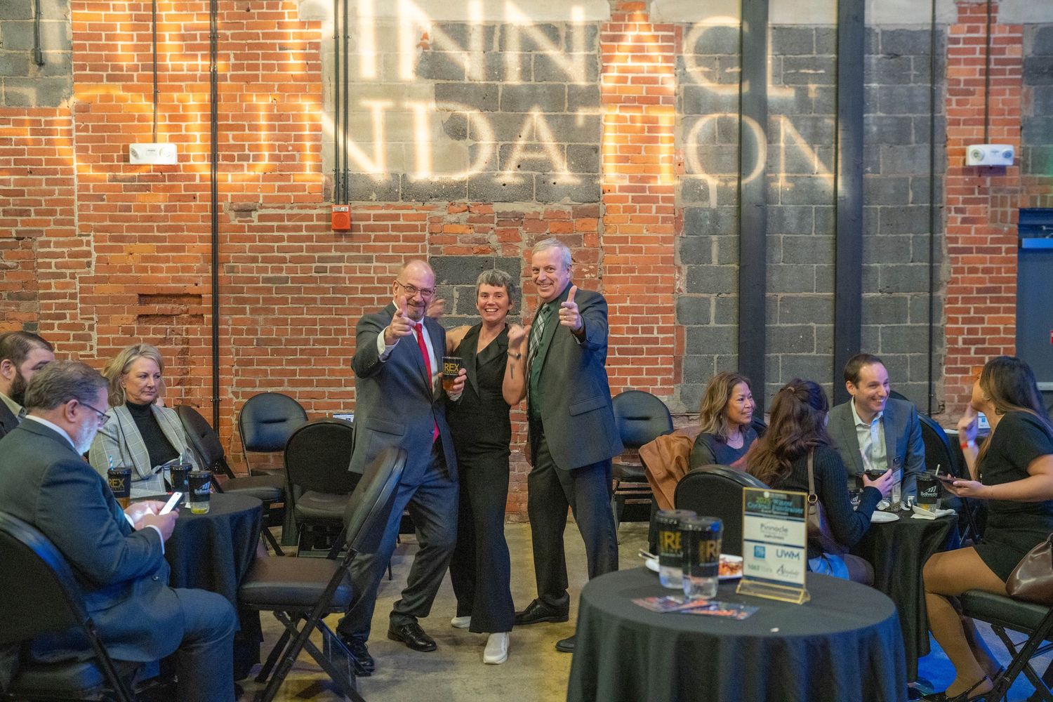 Three people pose and smile at a networking event inside a brick-walled venue with the Pinnacle Foundation sign behind them.