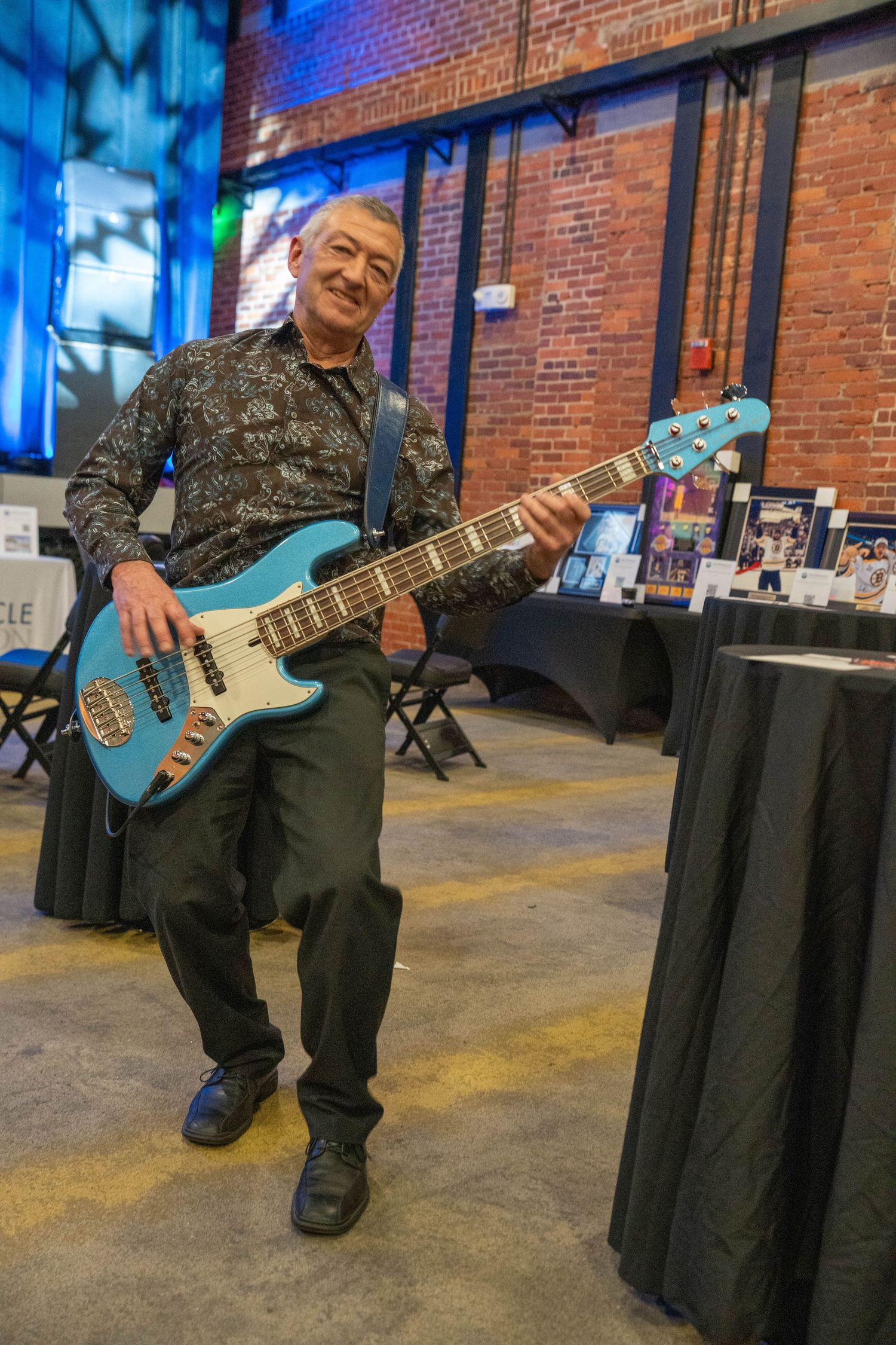 Smiling man sitting and playing a teal electric bass guitar at an indoor music event with brick walls.