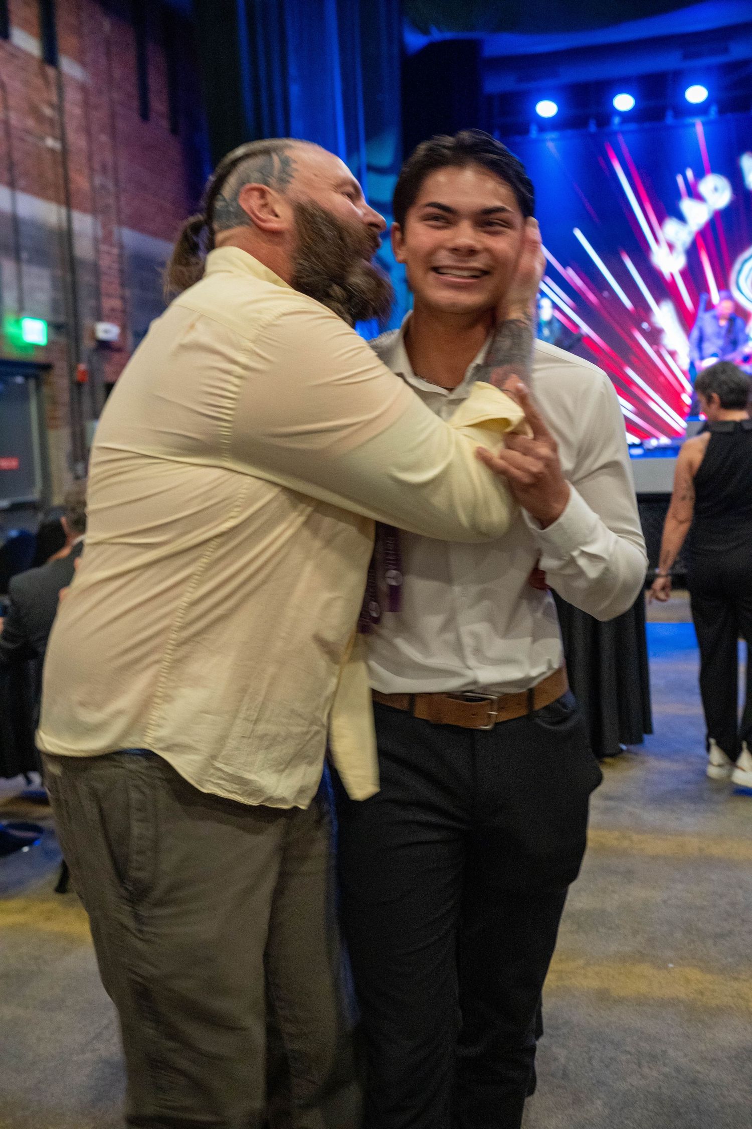 Two men sharing a joyful embrace at an event venue with colorful stage lighting in the background.