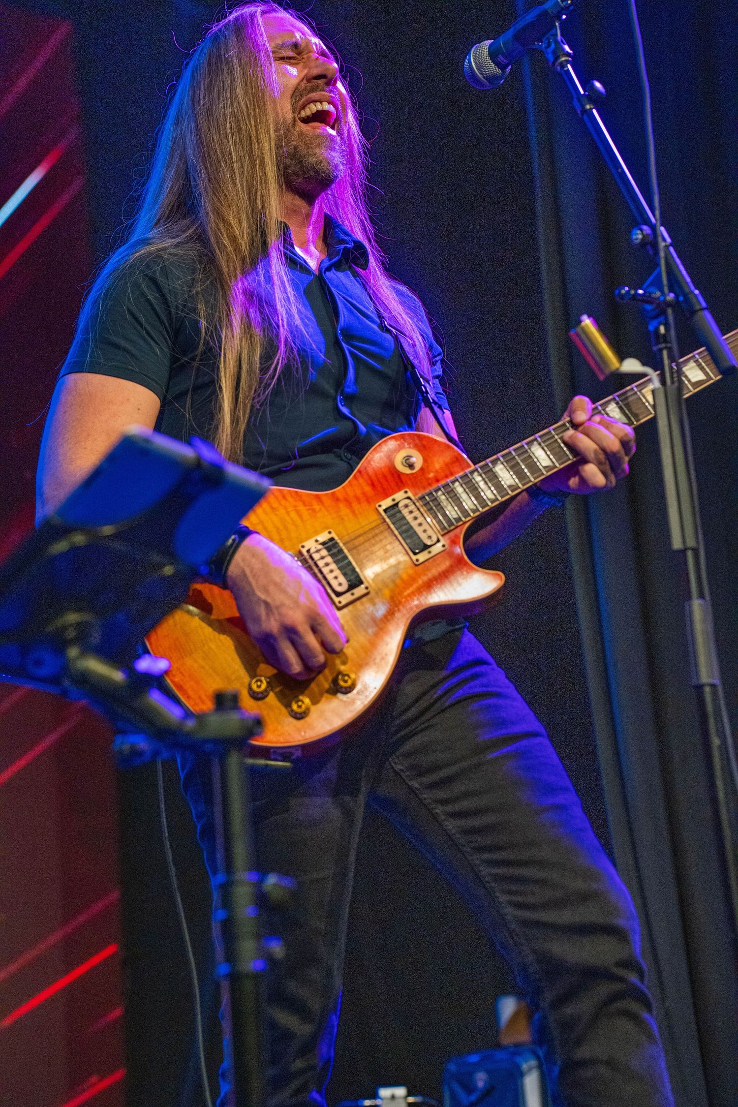 Long-haired male guitarist playing a sunburst Les Paul electric guitar on stage under colorful purple and blue lights.