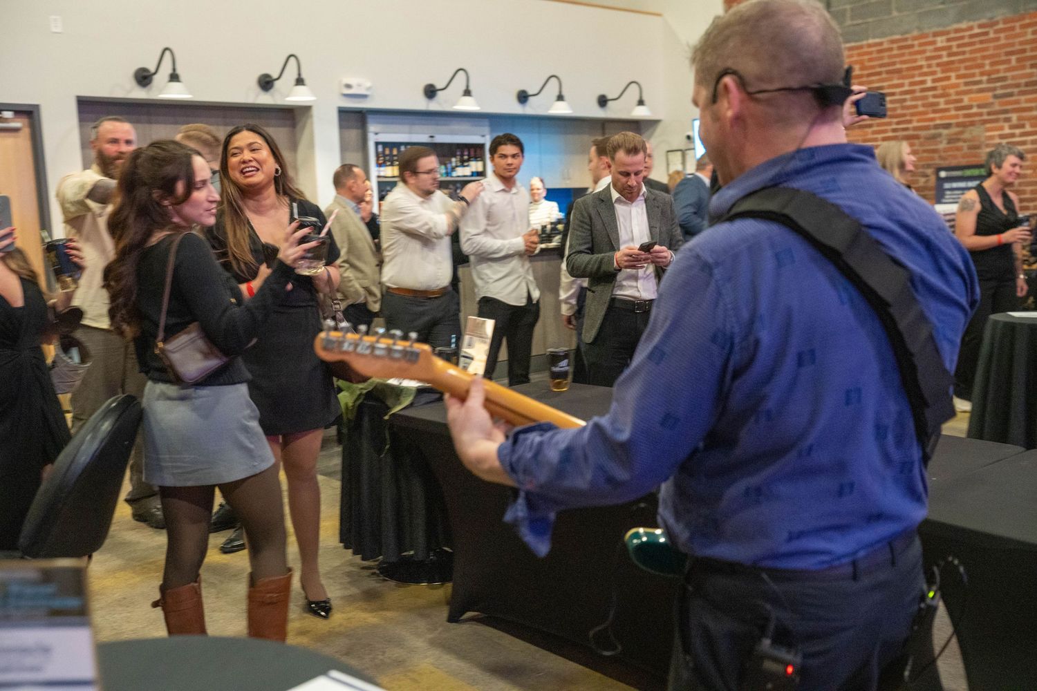 Guitarist performs for a crowd at an indoor event, with guests mingling and enjoying drinks in the background.