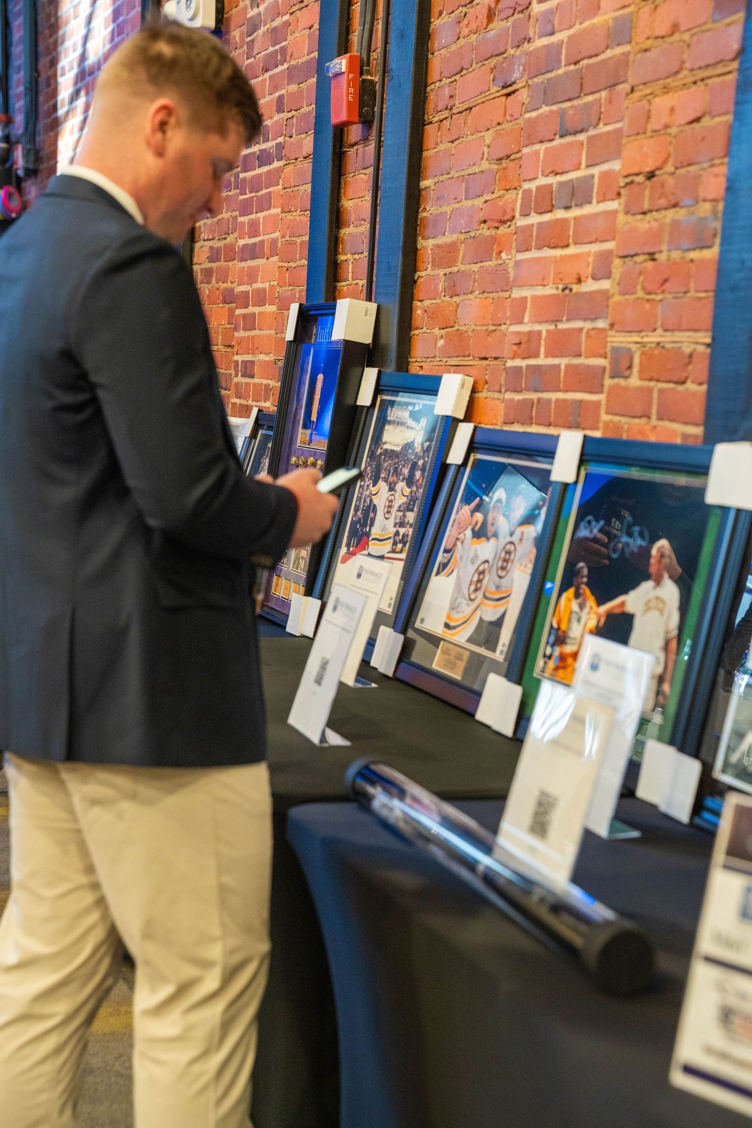 Man in blazer browsing framed sports memorabilia at a silent auction event near a brick wall.