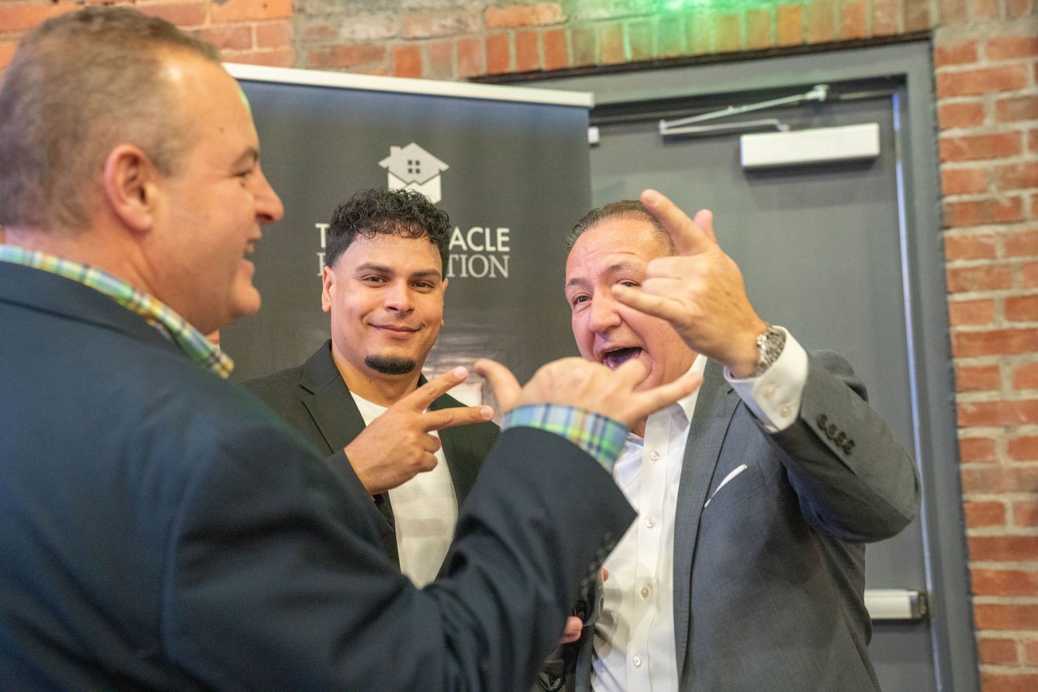 Three men laughing and making hand gestures at a Pinnacle Foundation event indoors.