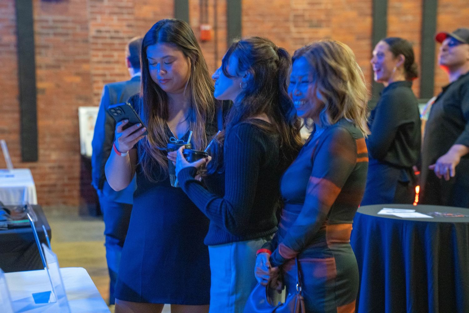 Three women gather closely together at an indoor event, looking at a smartphone in a dimly lit venue.