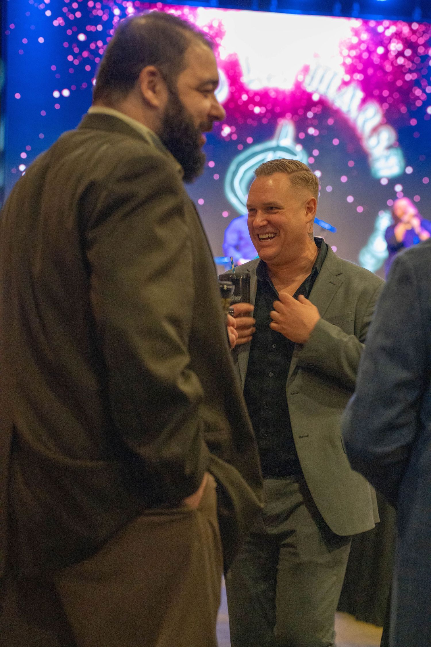 Two men in blazers laughing and conversing at a colorful evening event with neon lights in background.