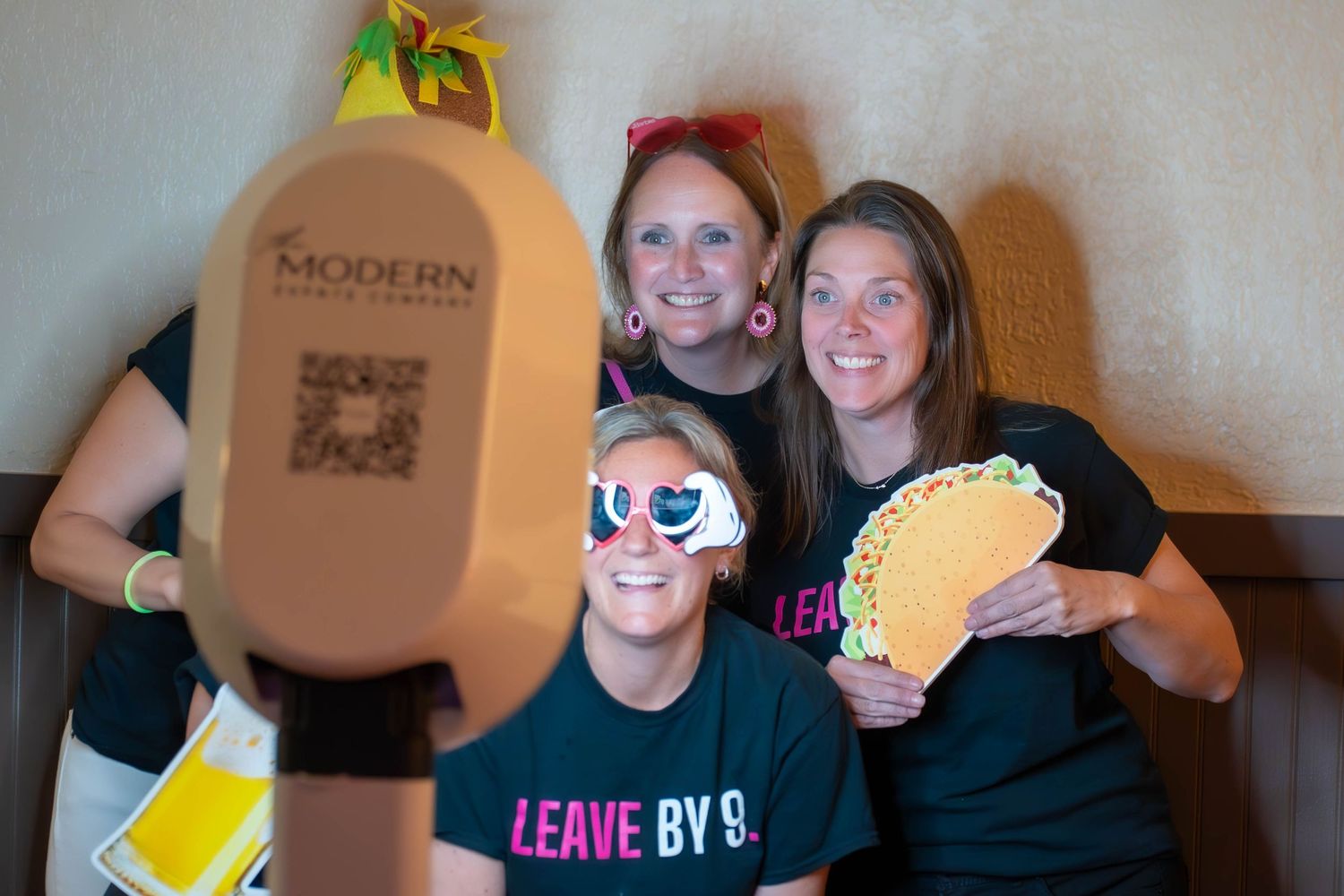 Three women posing at a photo booth with fun props including taco and novelty sunglasses at Modern Event Venue.