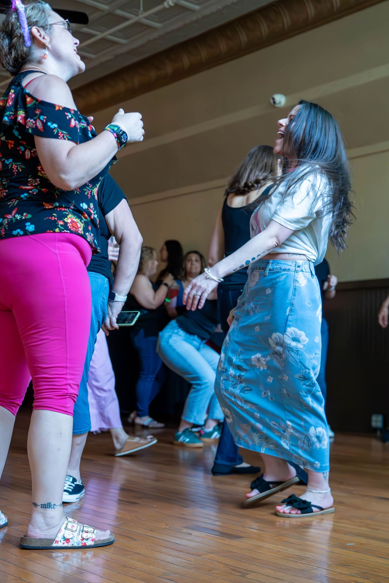 Women dancing joyfully together on a wooden dance floor at an indoor social dance event.