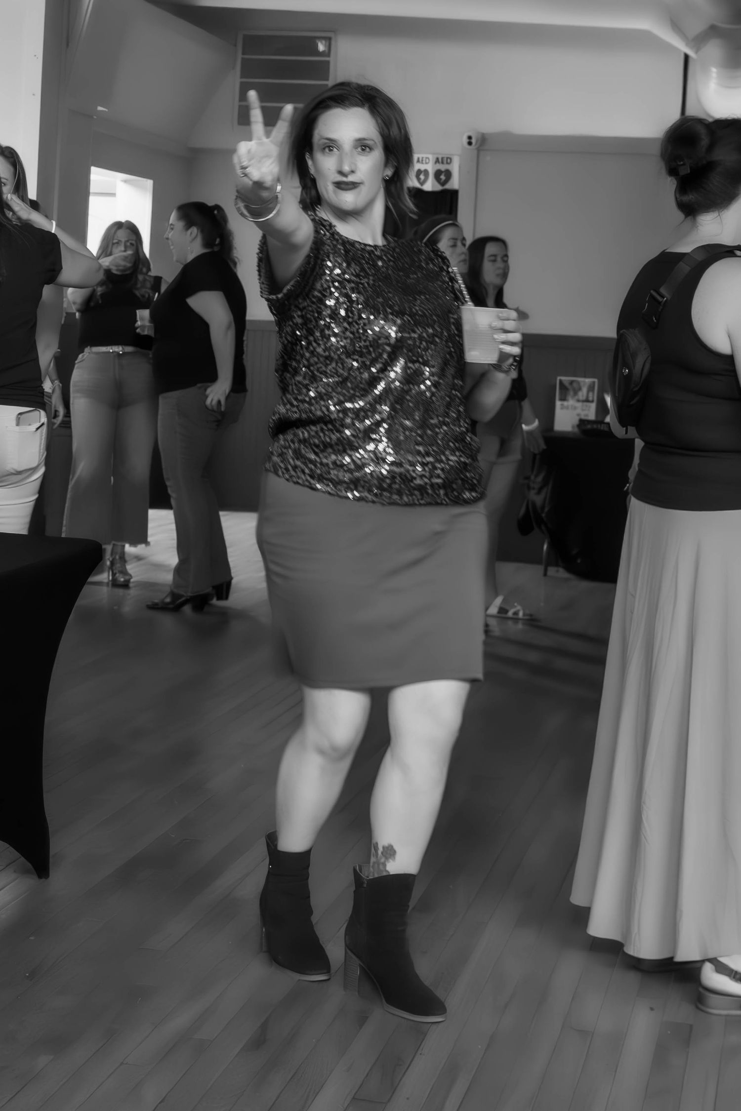 Woman in sequin top and skirt flashing peace sign while holding drink at indoor social gathering, black and white photo.