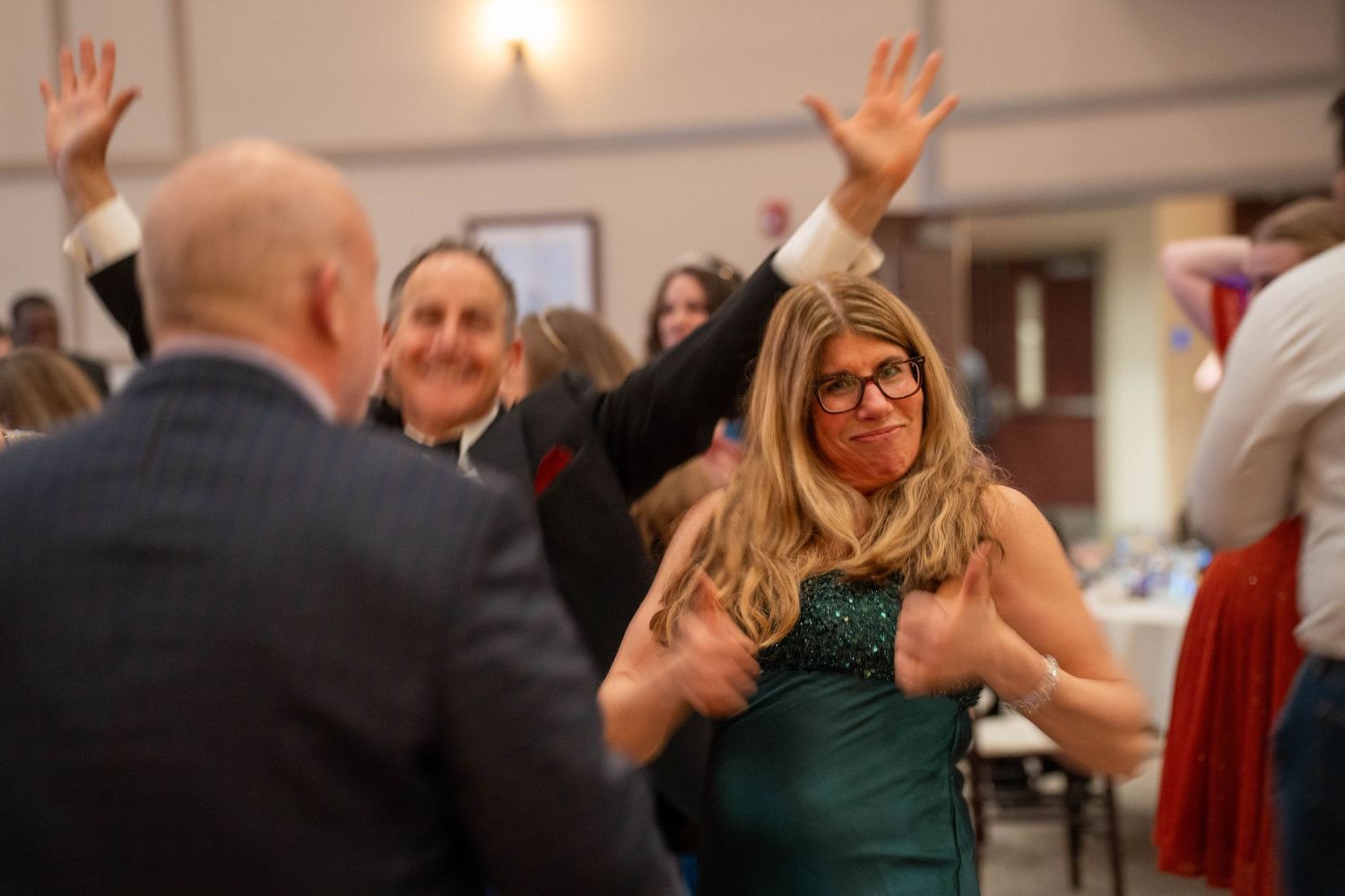 People dancing and celebrating at a formal event, woman in green dress smiling with hands raised in a festive hall.