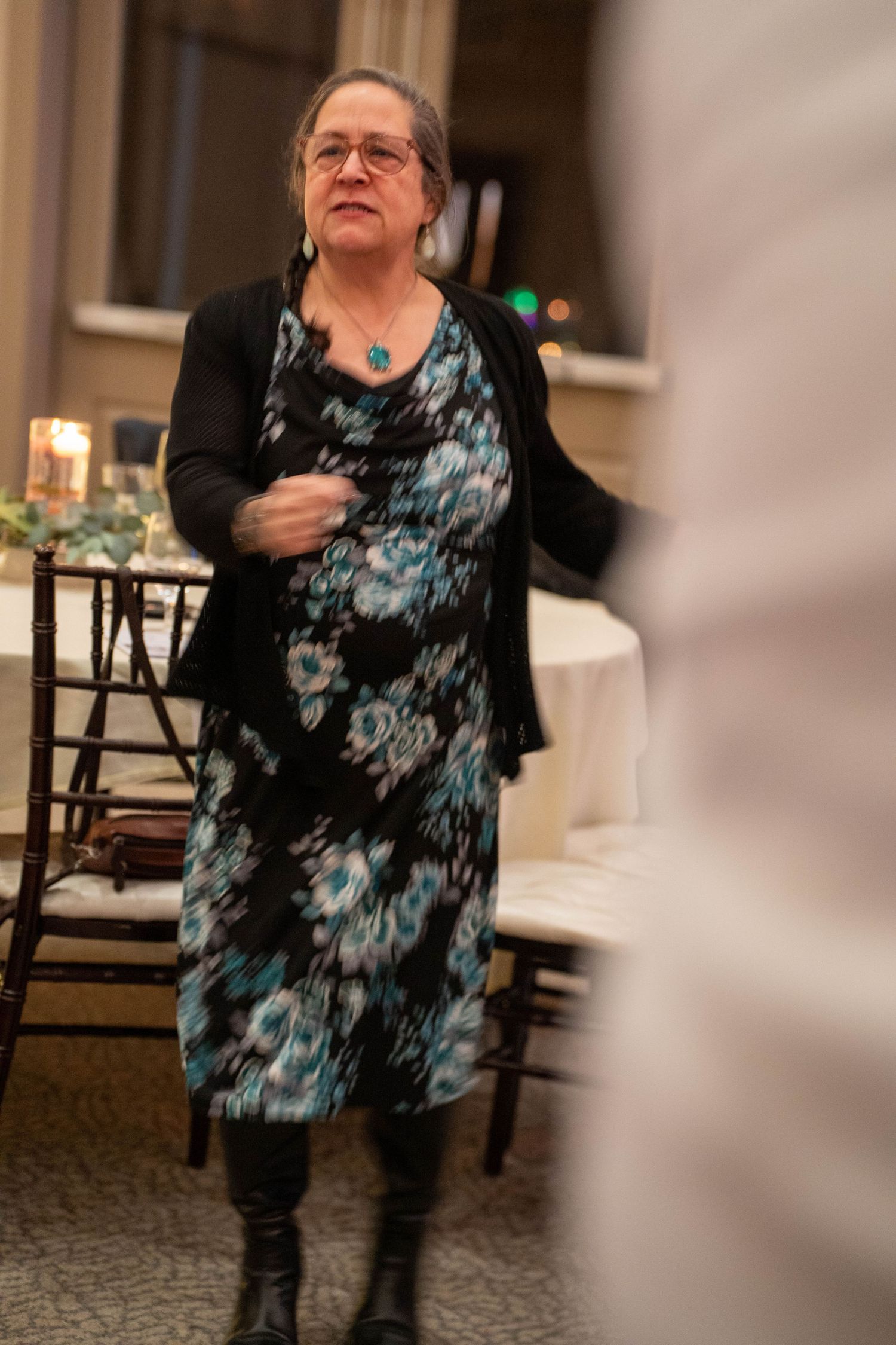 Woman in floral dress and black cardigan with turquoise necklace standing at an elegant indoor event.