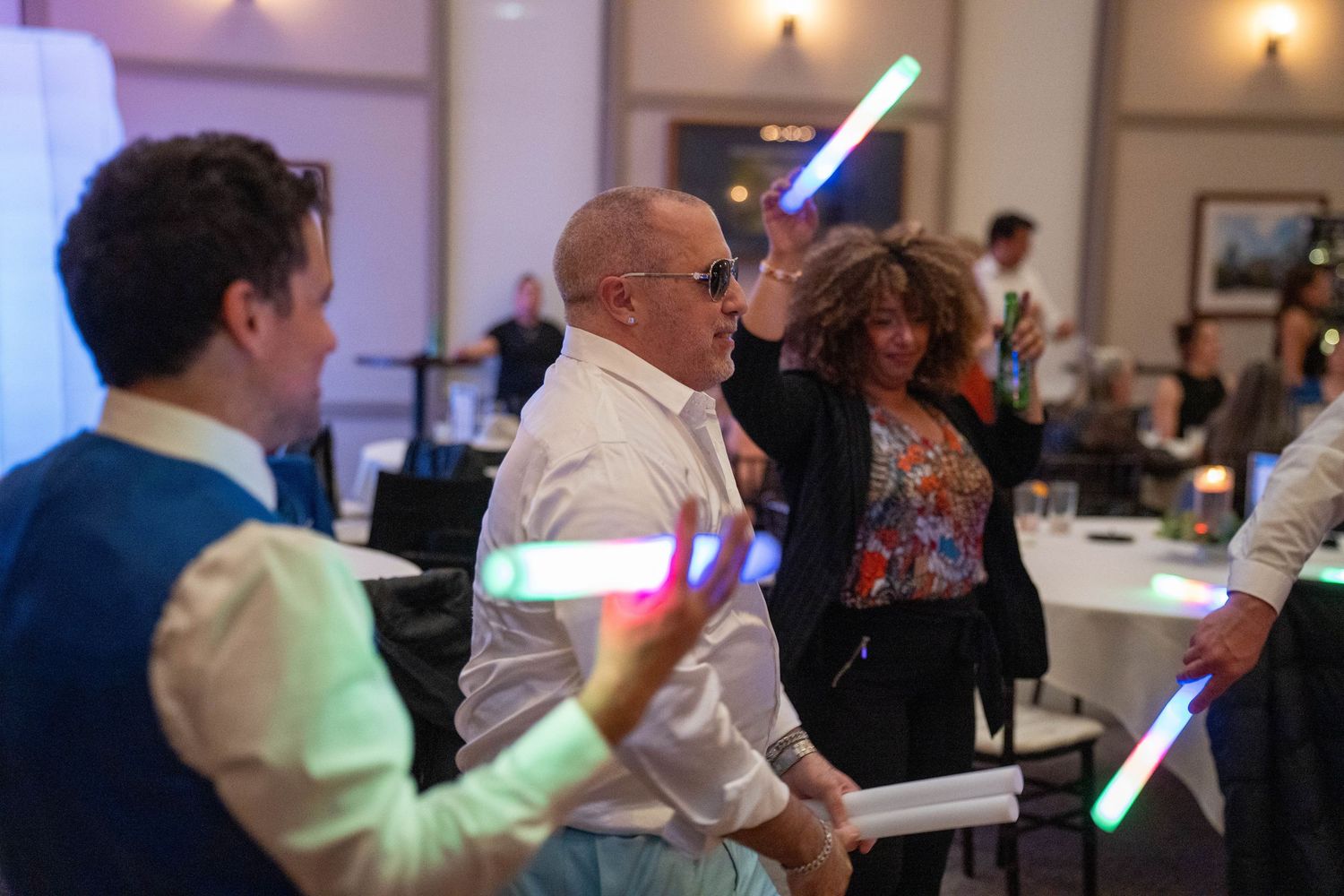 Guests dancing and waving colorful LED glow sticks at a lively indoor party or wedding reception event.