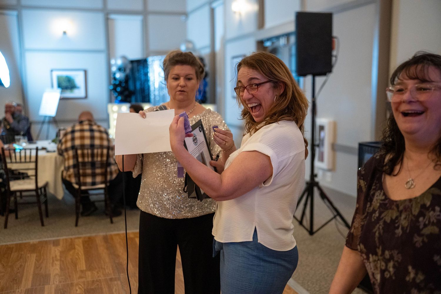Women laughing and celebrating at an indoor event, one holding a card and gift at a festive gathering.
