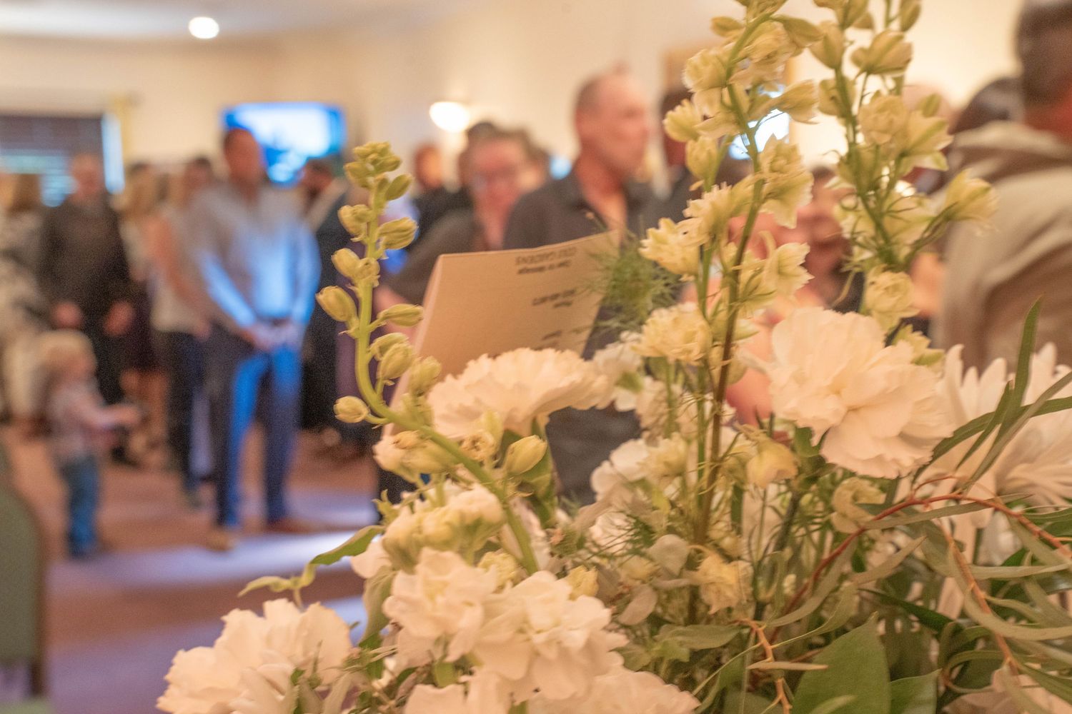 White flowers in foreground with blurred crowd of people gathered indoors at a formal event.