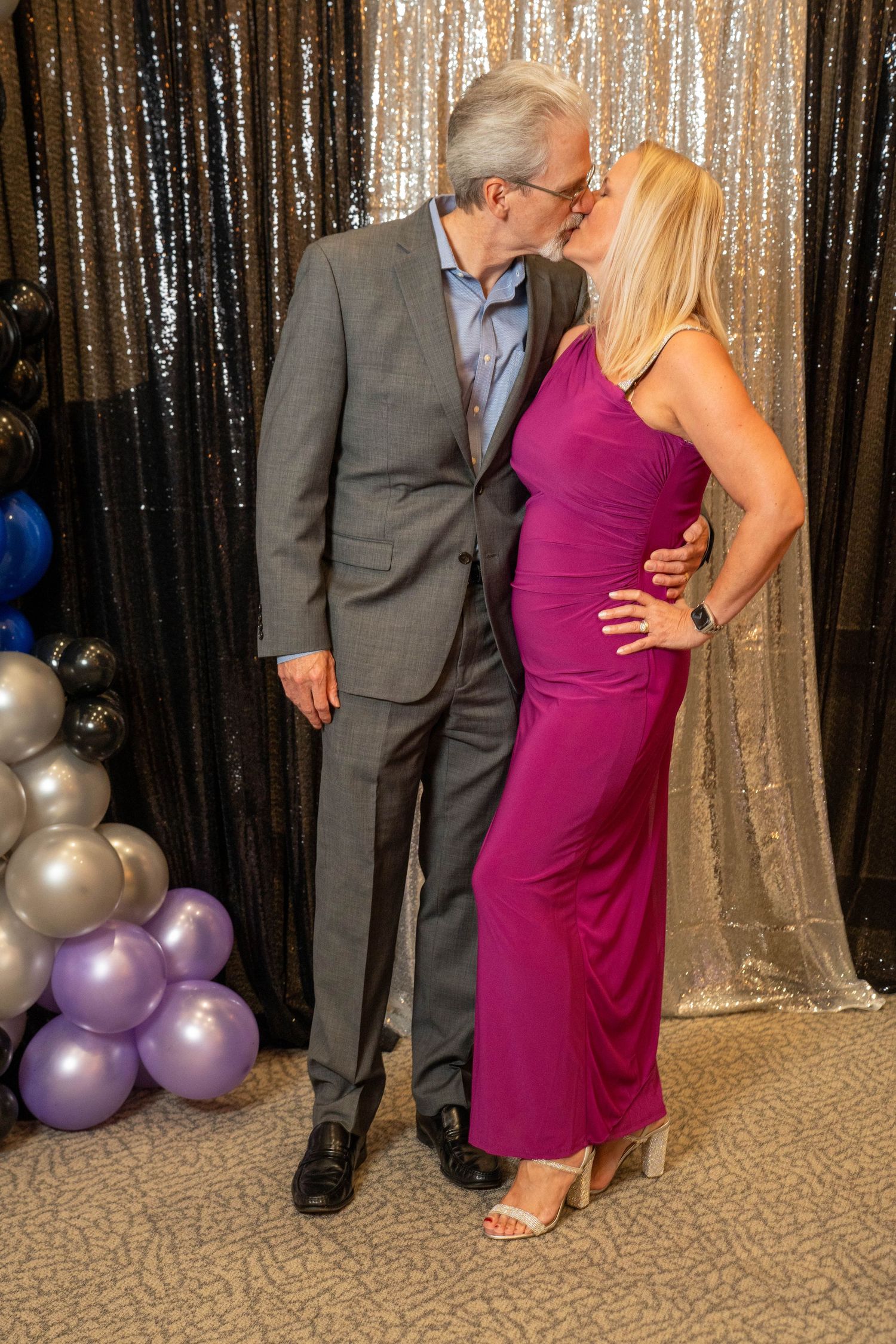 Couple kissing at formal event, man in gray suit and woman in magenta gown, sequin backdrop with balloons.