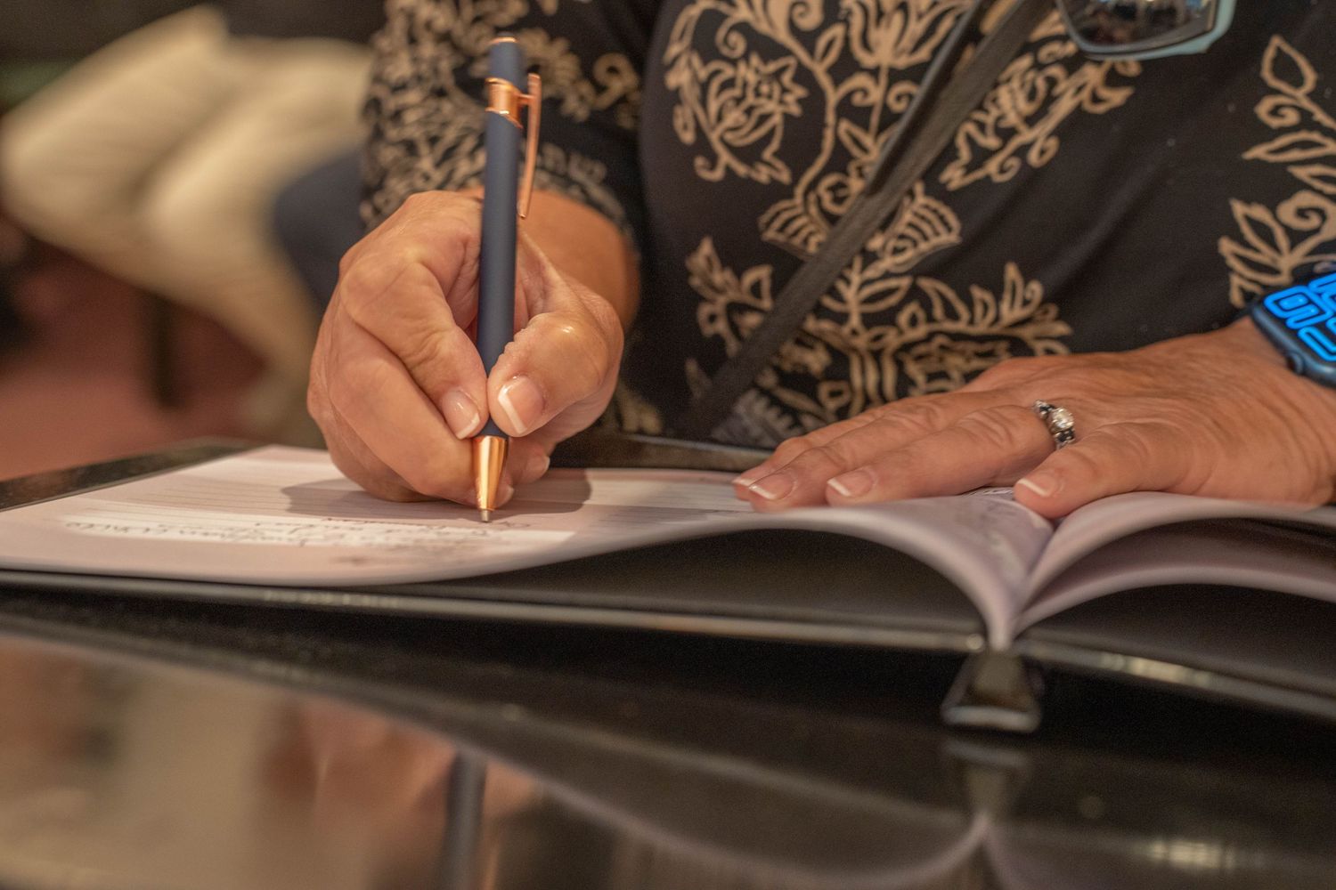 A person wearing a patterned shirt writes in an open book with a pencil, wearing a ring and blue bracelet.