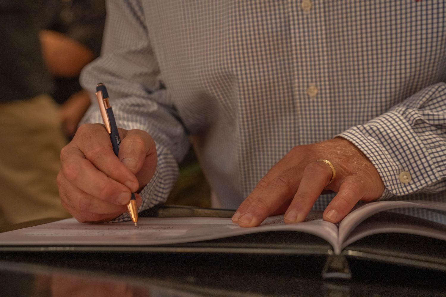 Man in checkered shirt writing in open notebook with pen, wearing gold wedding ring.