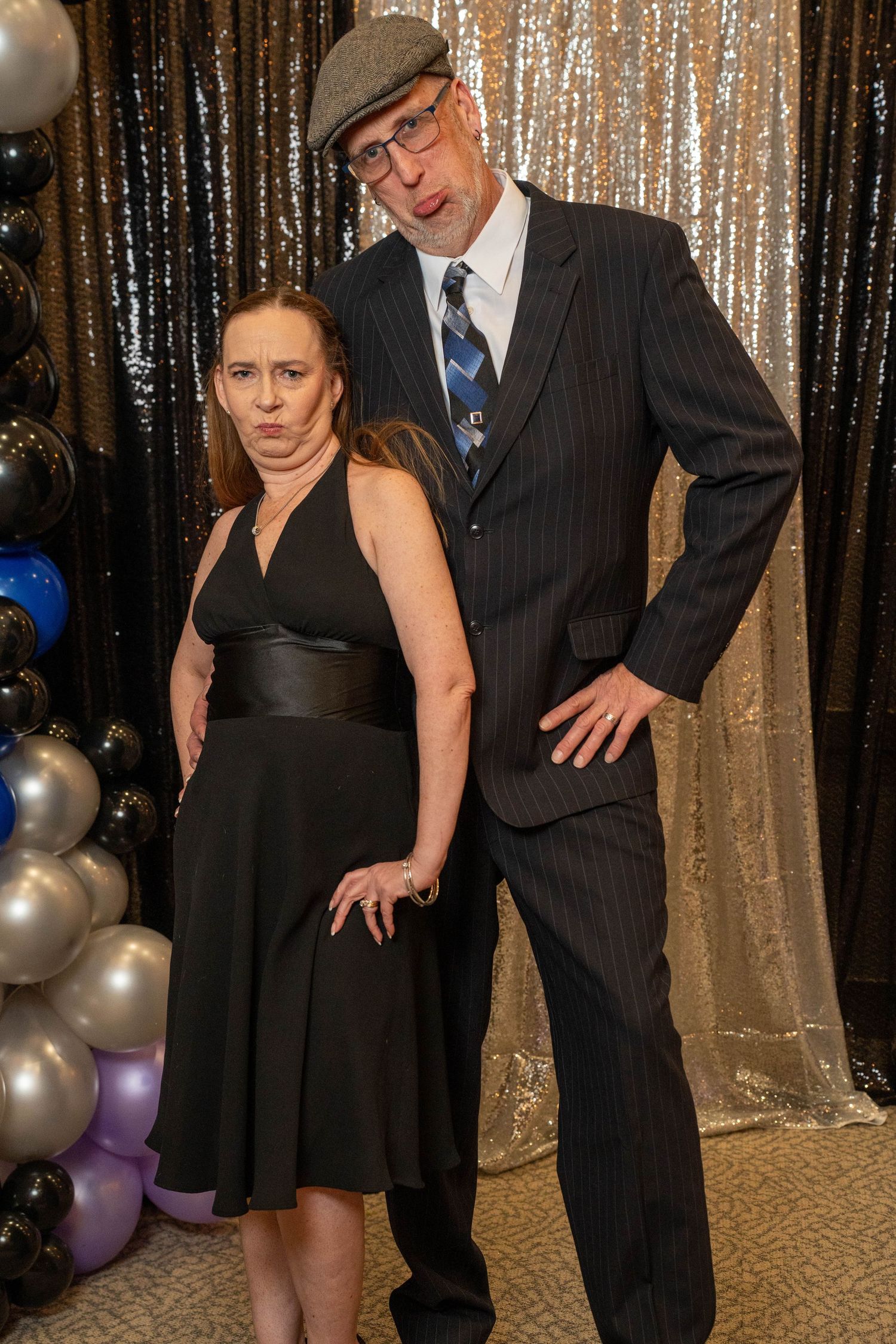 Couple posing formally at a gala event, woman in black dress, man in pinstripe suit and flat cap, sequin backdrop.