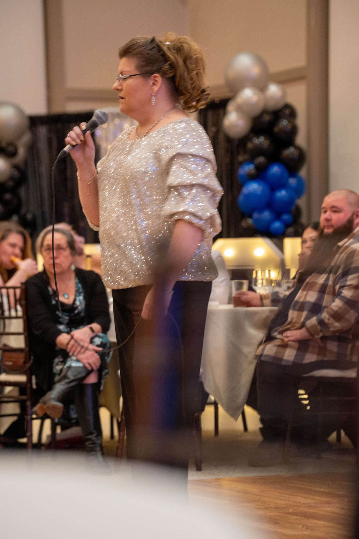 Woman in sequin top speaking at microphone during formal event with black and blue balloon decorations.