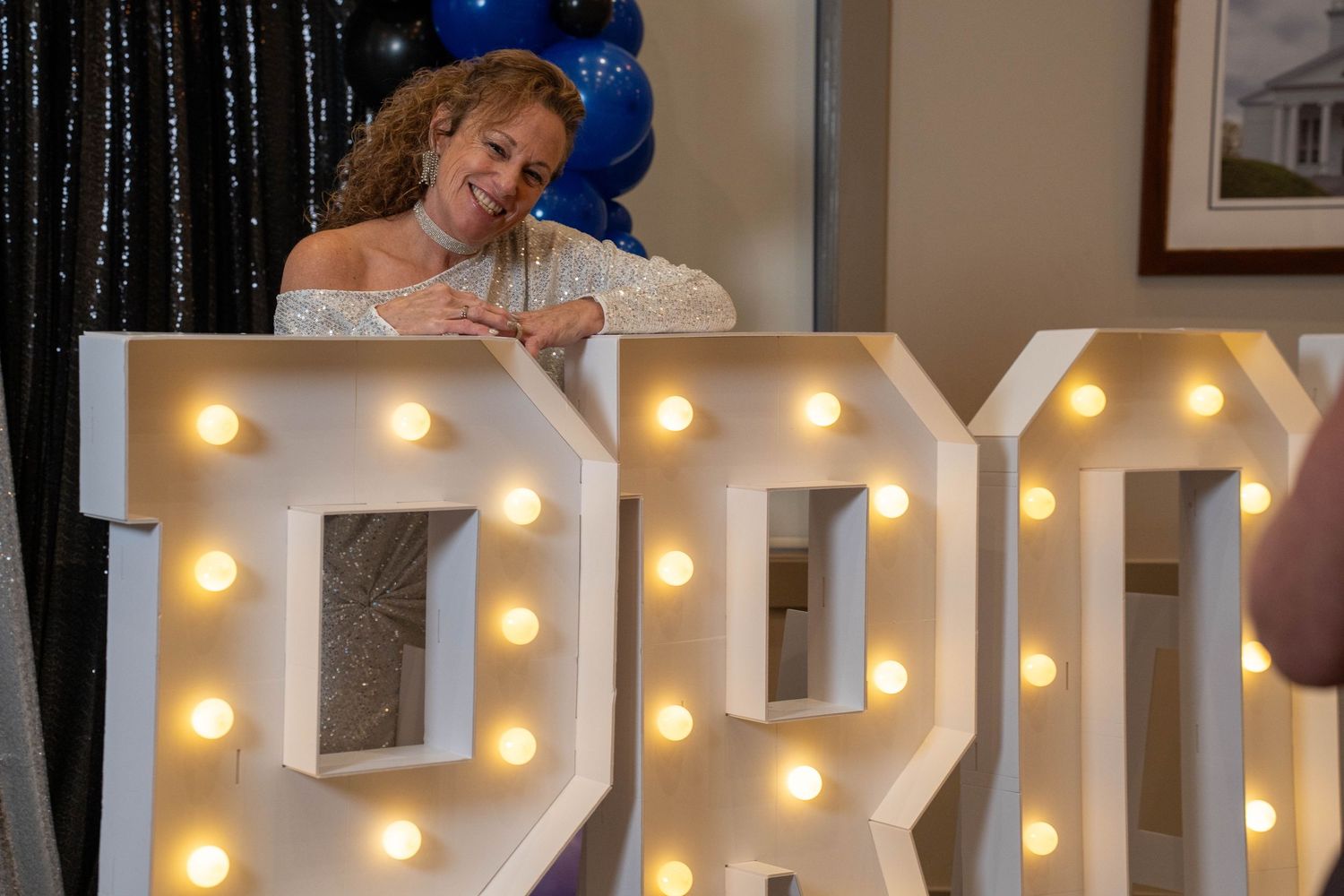 Woman posing with illuminated marquee letters spelling PRO at a prom event with blue balloons.