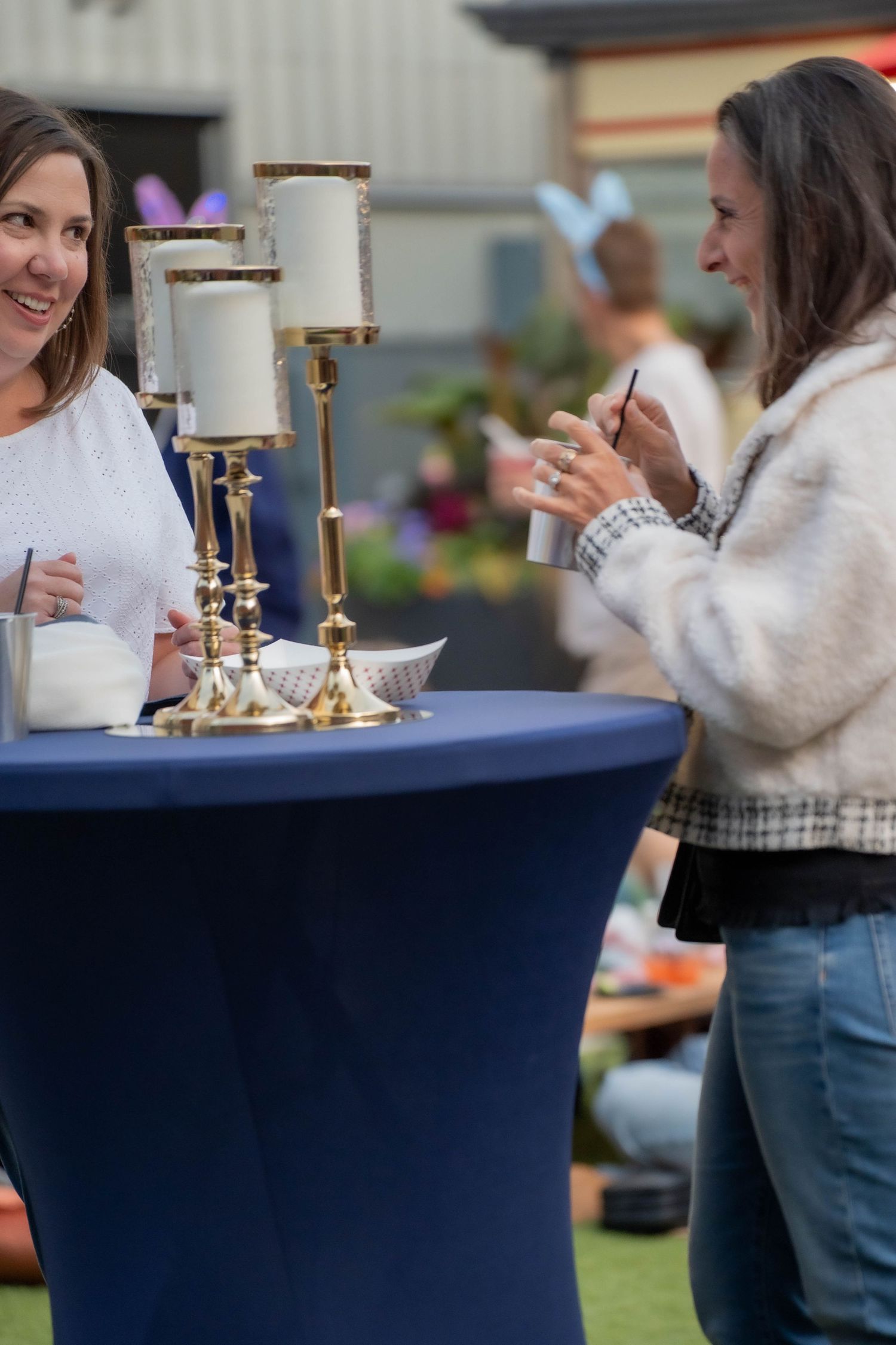 Two women chat at a blue cocktail table with gold candle holders at an outdoor event.