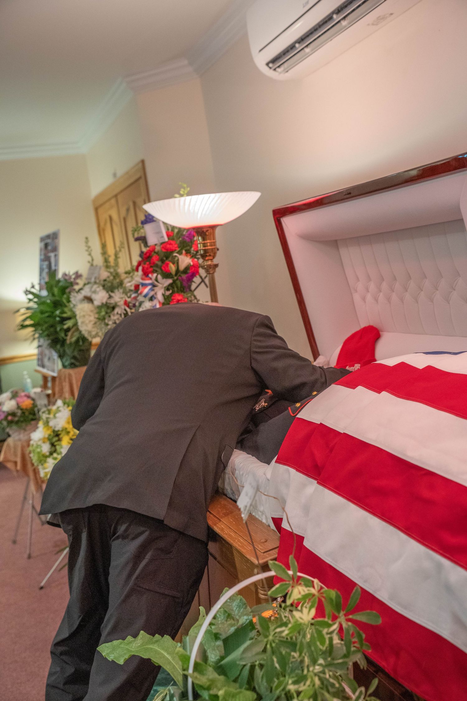 Man in dark suit leaning over open casket draped with American flag at funeral home with floral arrangements.