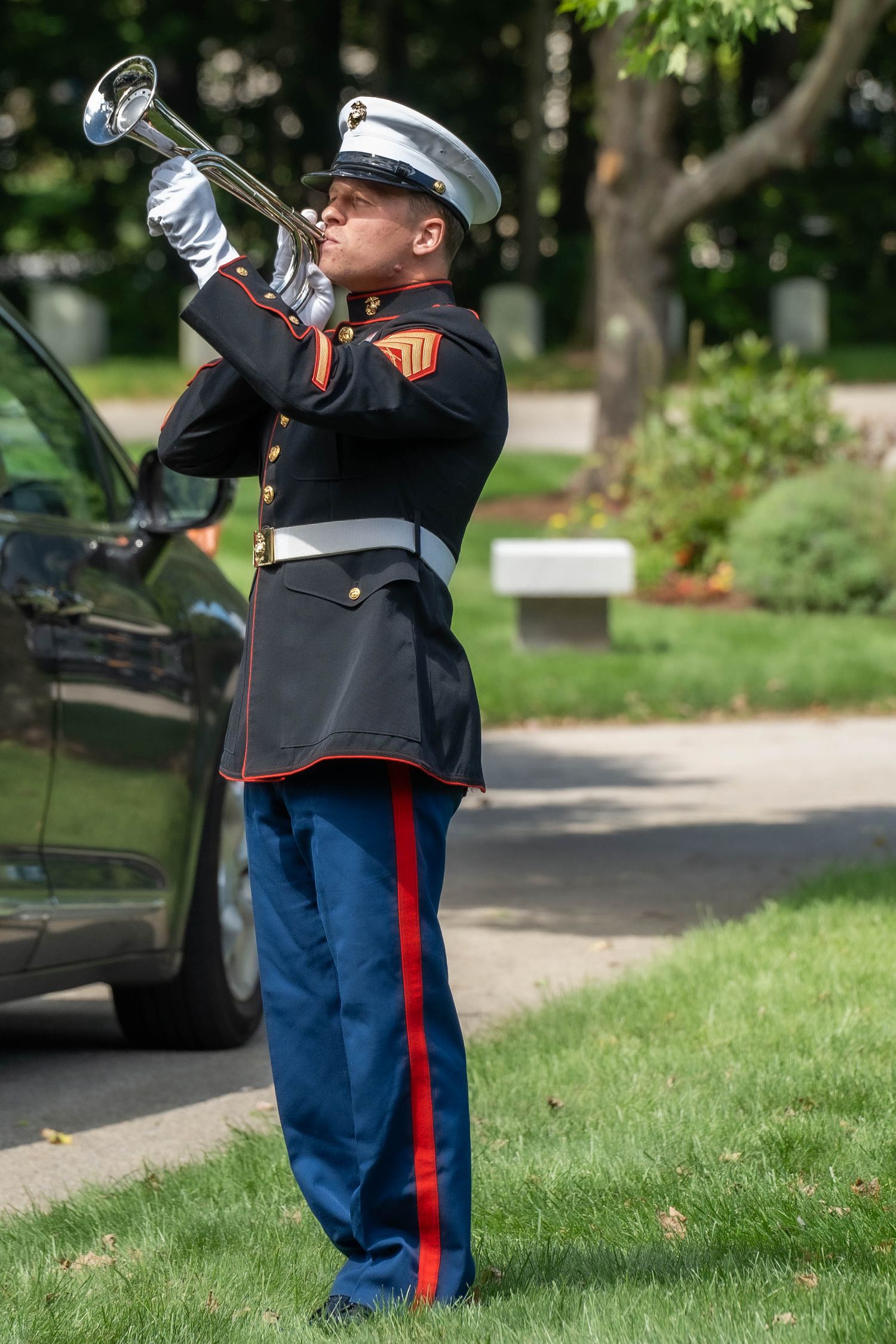 US Marine in dress uniform playing a bugle at a military funeral ceremony in a cemetery.