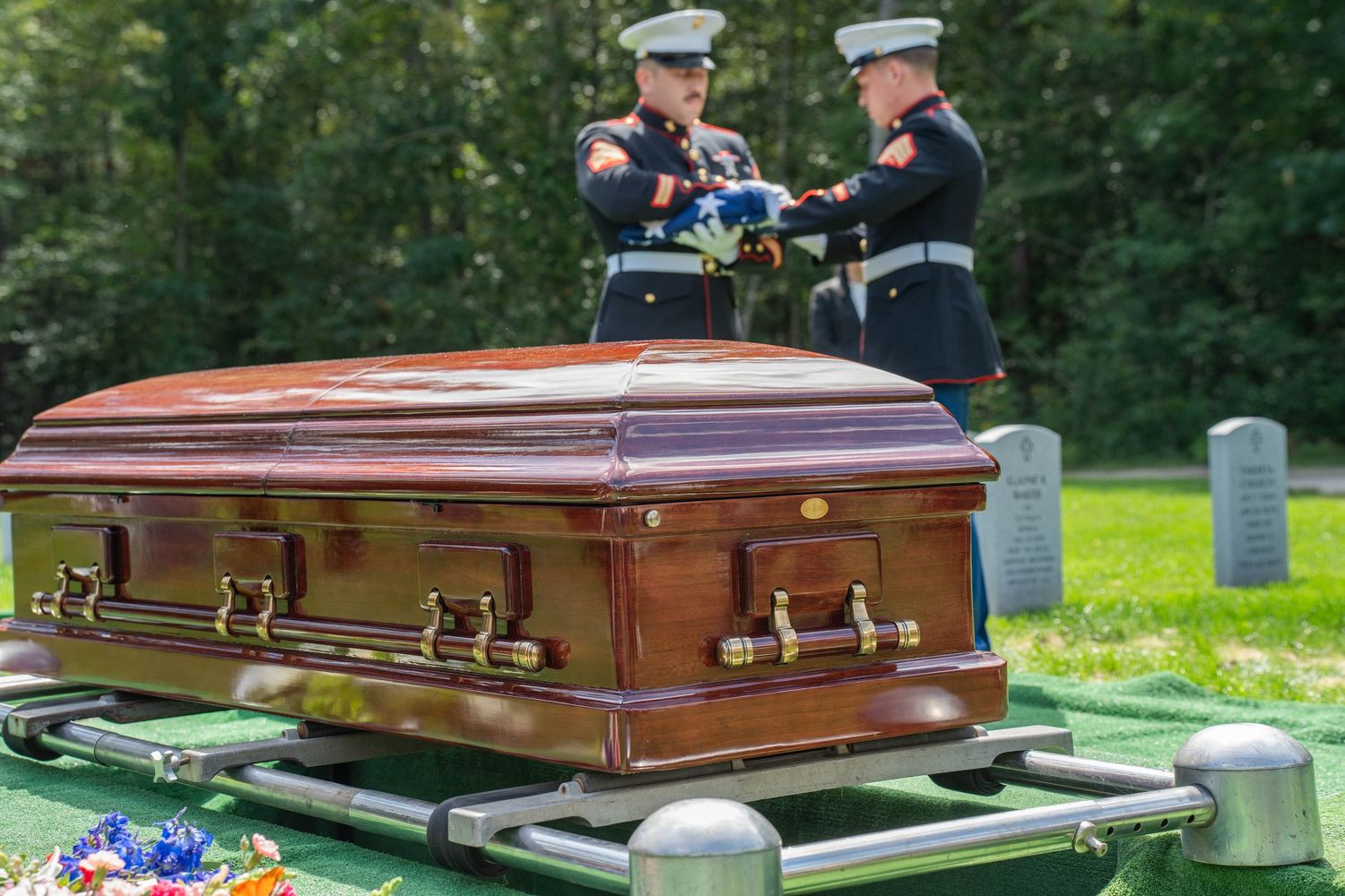 Two Marines fold an American flag over a wooden casket during a military funeral ceremony outdoors.