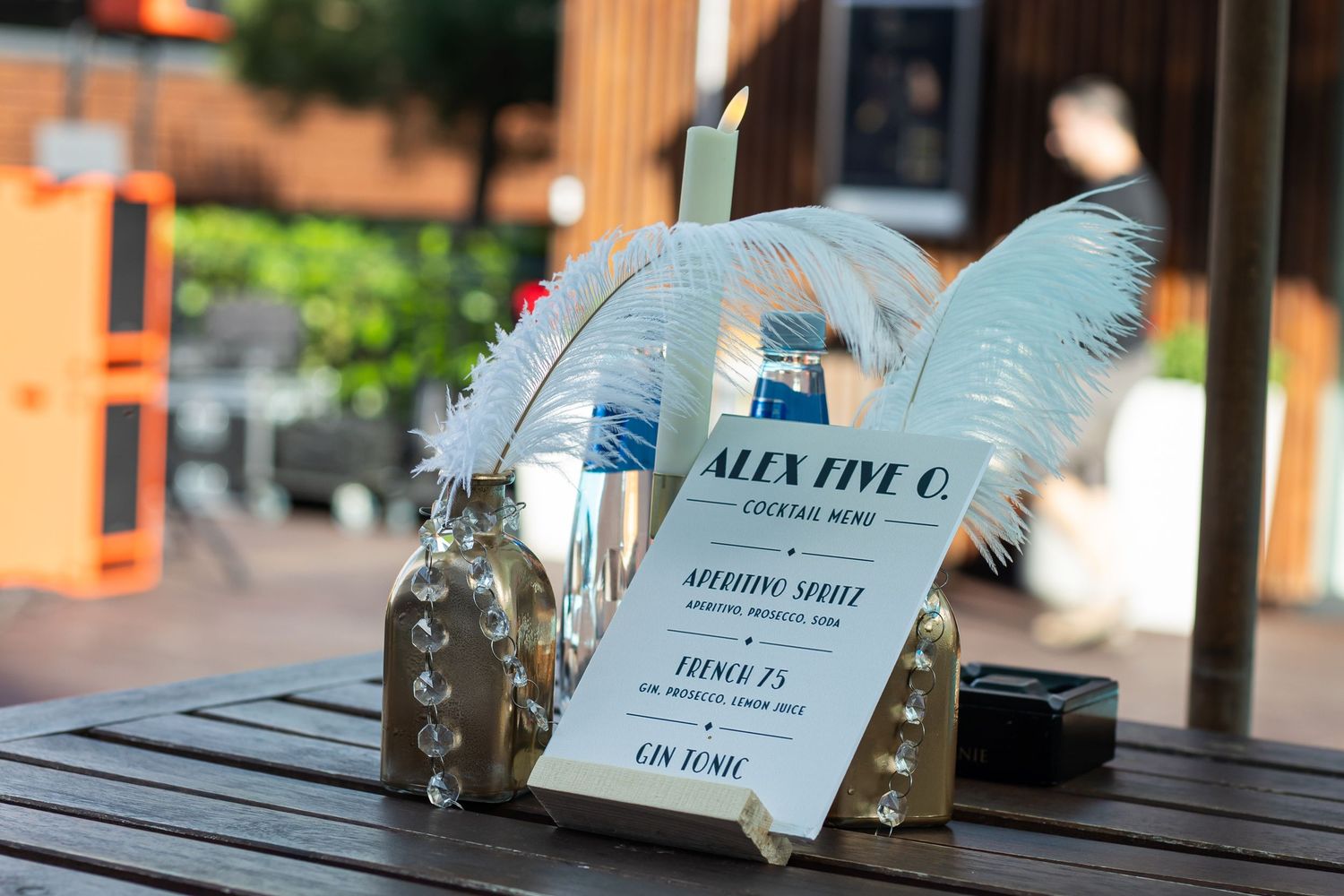 Elegant cocktail menu display with white feathers and glass bottles on a wooden table at Alex Five O bar.
