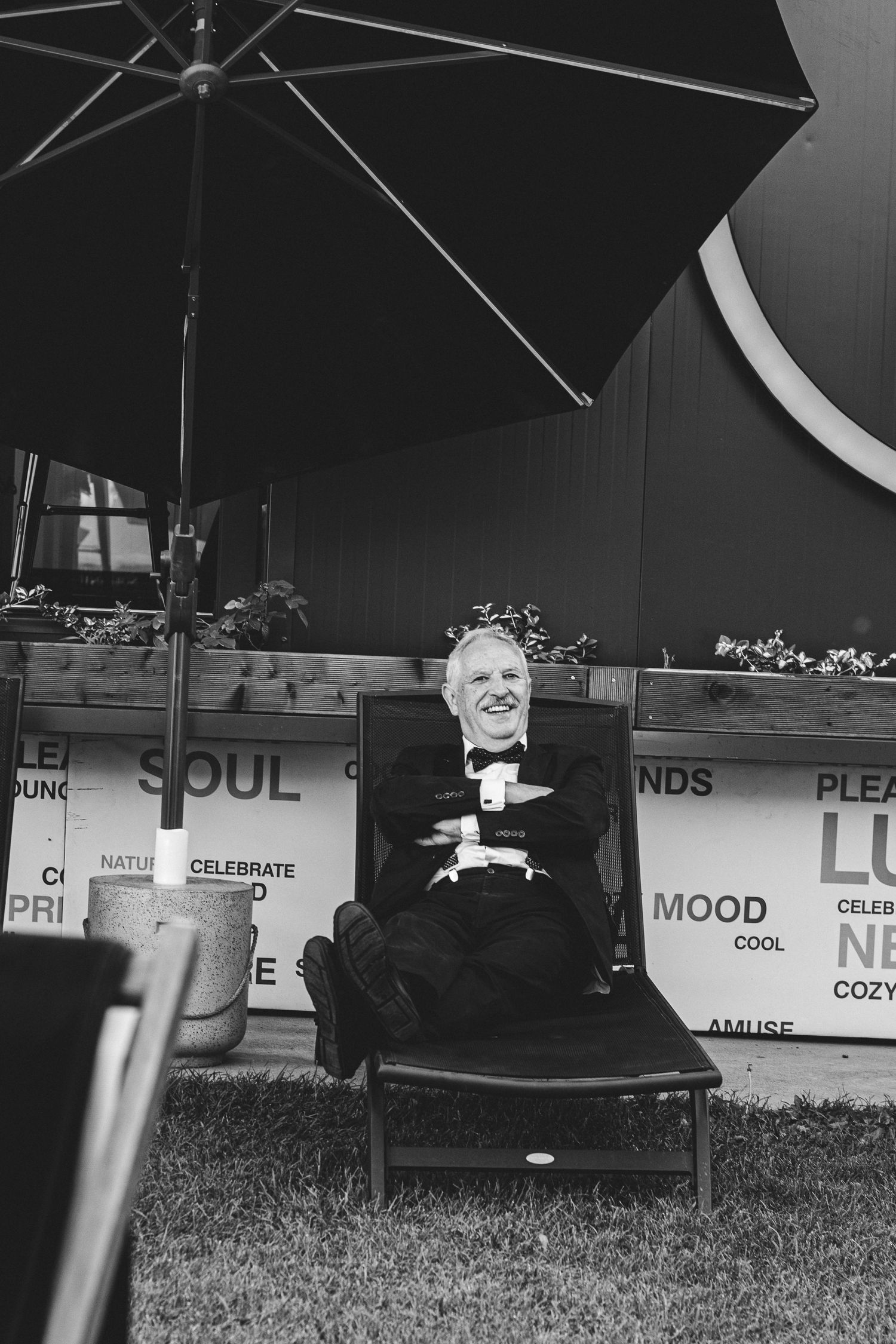 Elderly man in tuxedo laughing joyfully on a chair under a patio umbrella in a black and white photo.