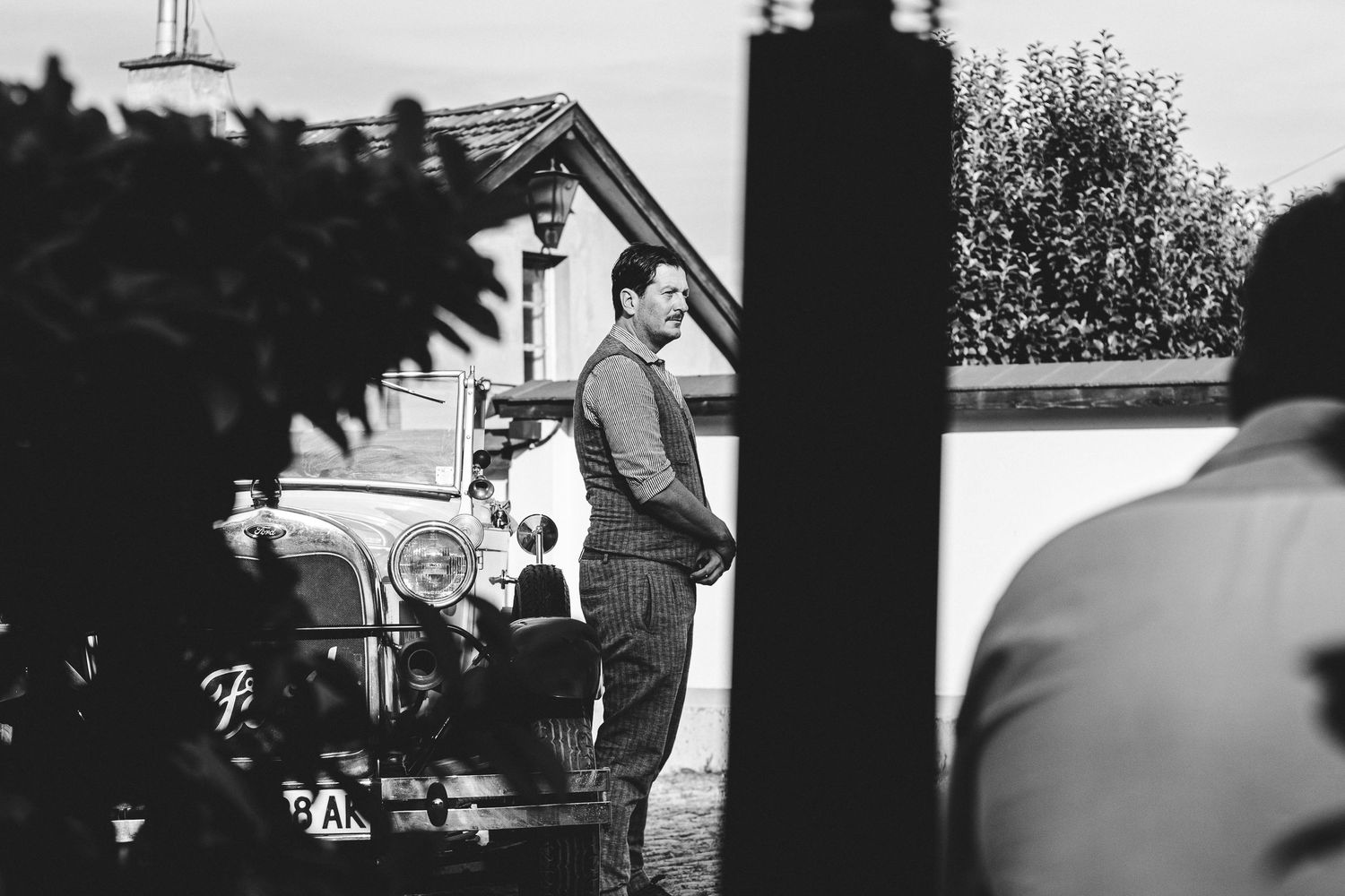 Black and white photo of a man standing beside a vintage classic car outside a historic building.