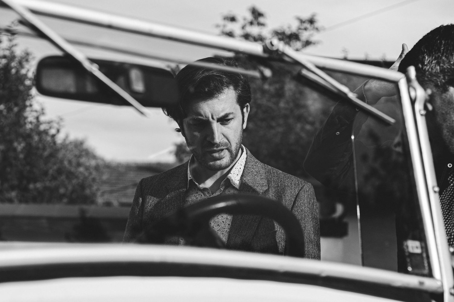 Black and white photo of a well-dressed man in a suit sitting in a vintage car, looking down thoughtfully.