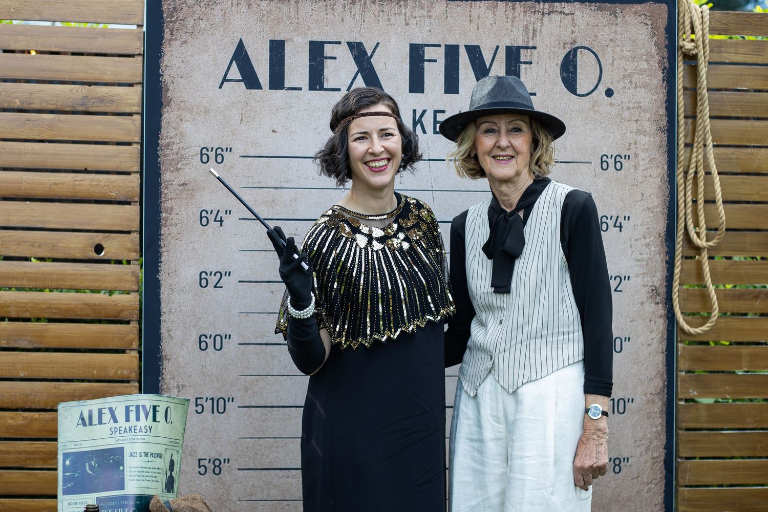 Two women posing in front of an Alex Five O. height chart backdrop, dressed in 1920s-style costumes.