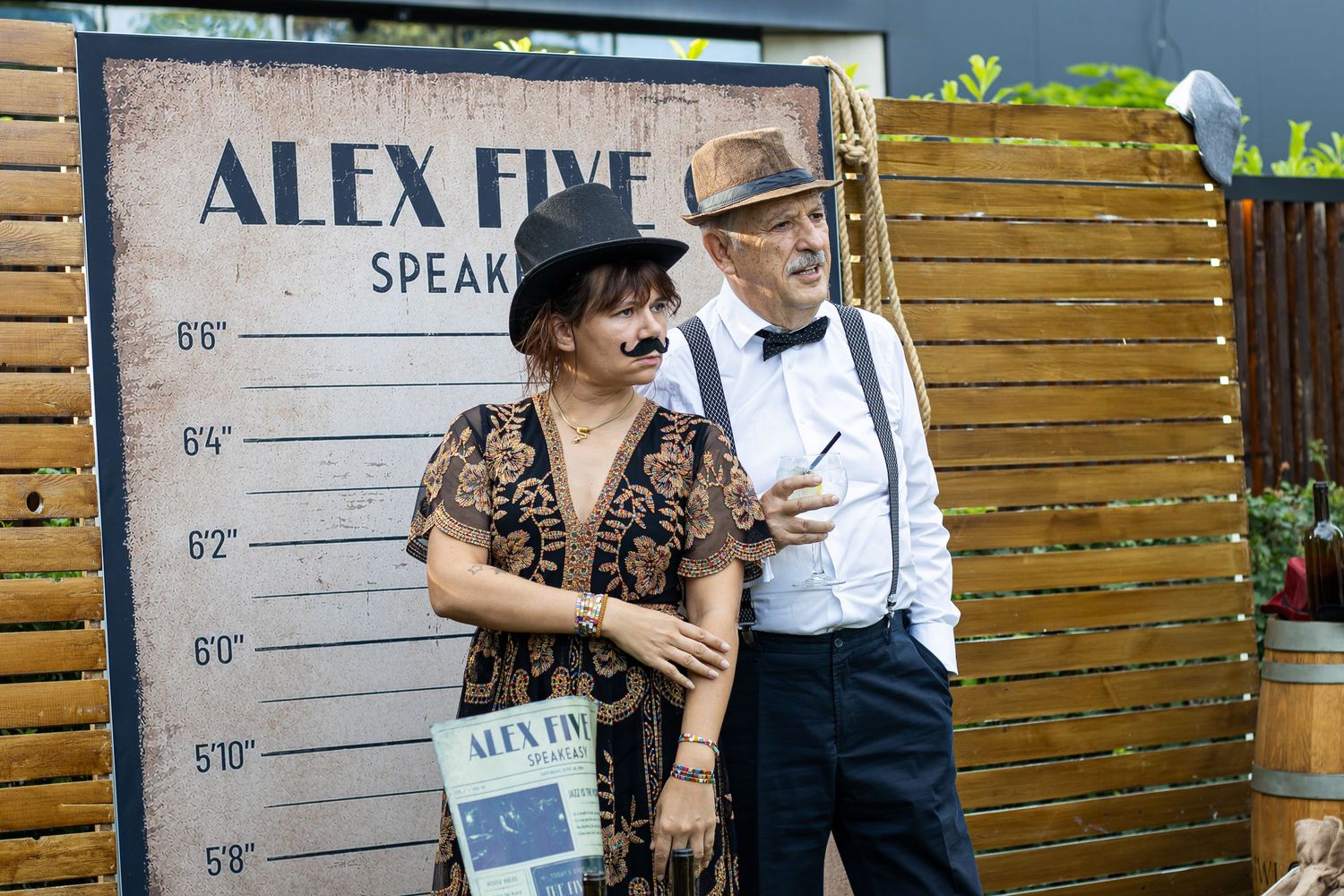 Two people in vintage costumes pose beside an Alex Five speaker event height chart backdrop outdoors.