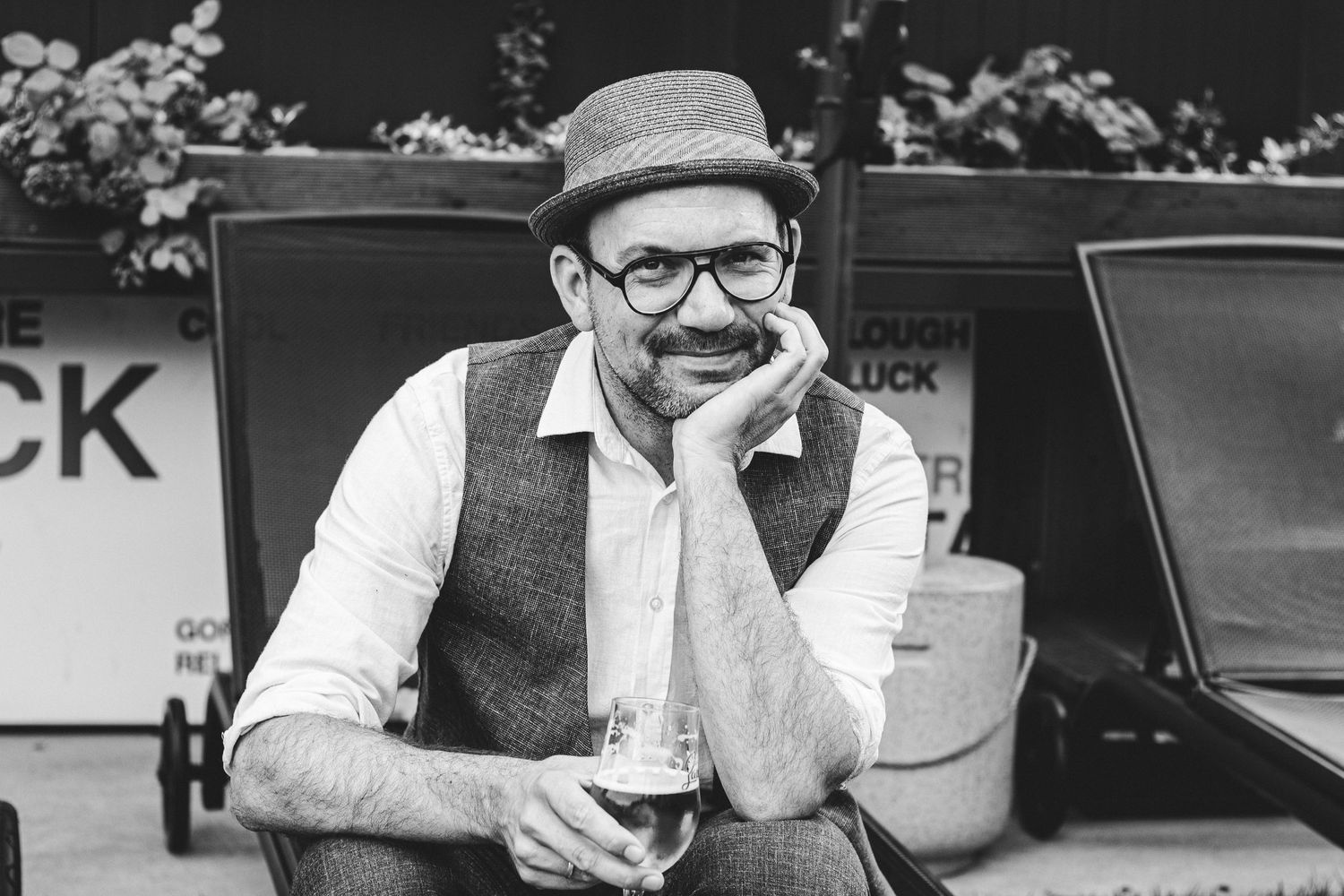 Black and white photo of a smiling bearded man in glasses, hat, and vest holding a drink outdoors.