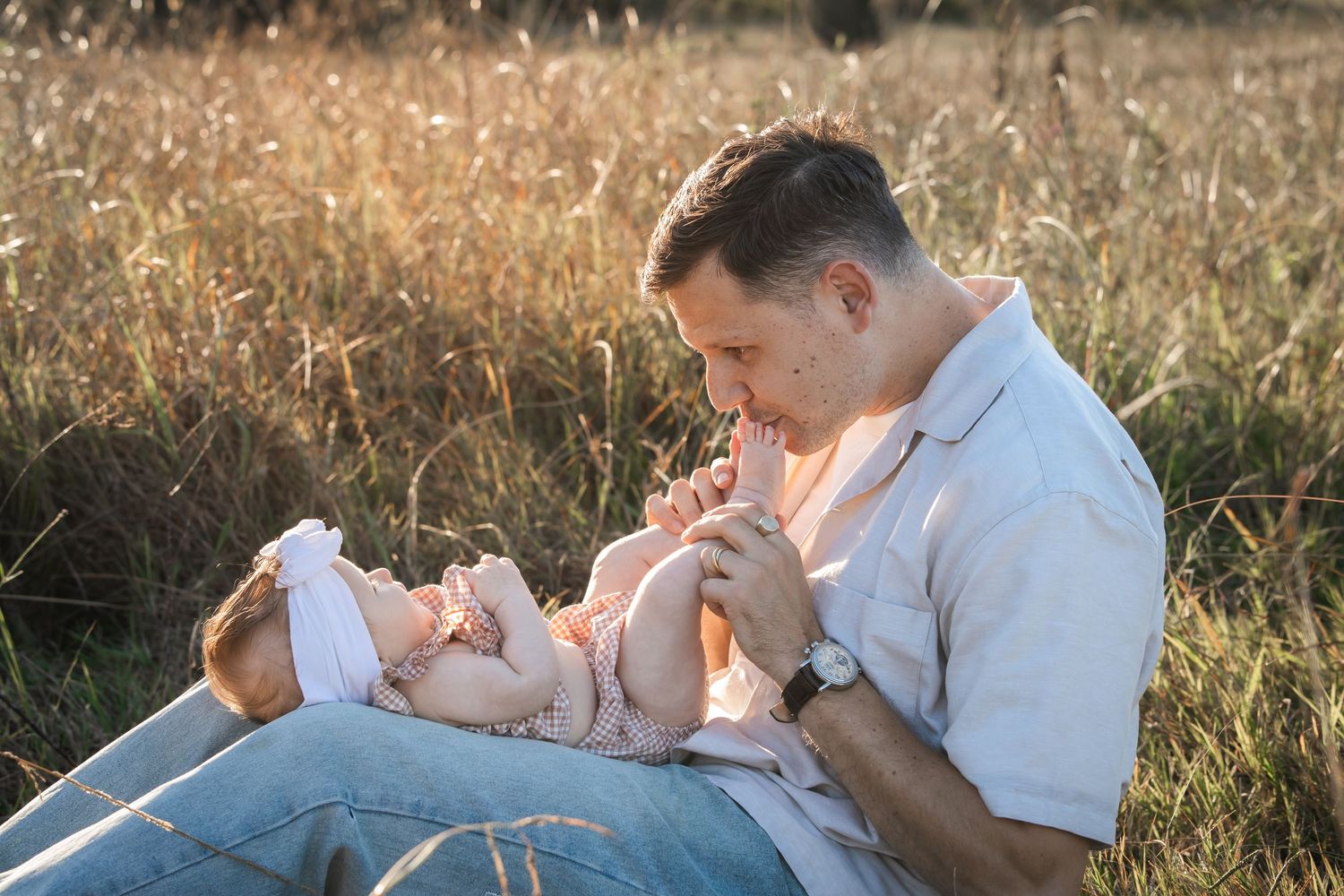 A father sits in a golden field, tenderly kissing his baby's feet during a sunset family photoshoot in Rouse Hill, Hills District Sydney