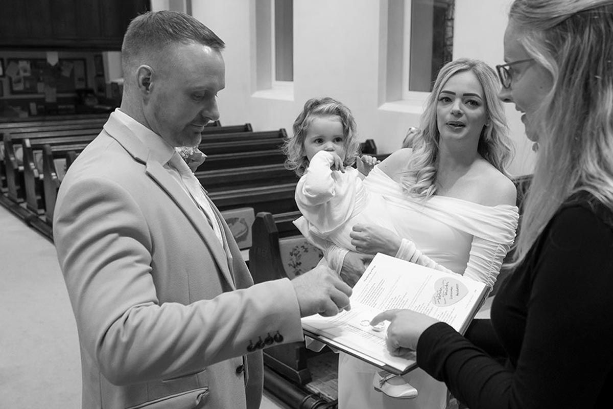 Black and white photo of a couple signing wedding documents in a church, officiant holding papers, child in background.