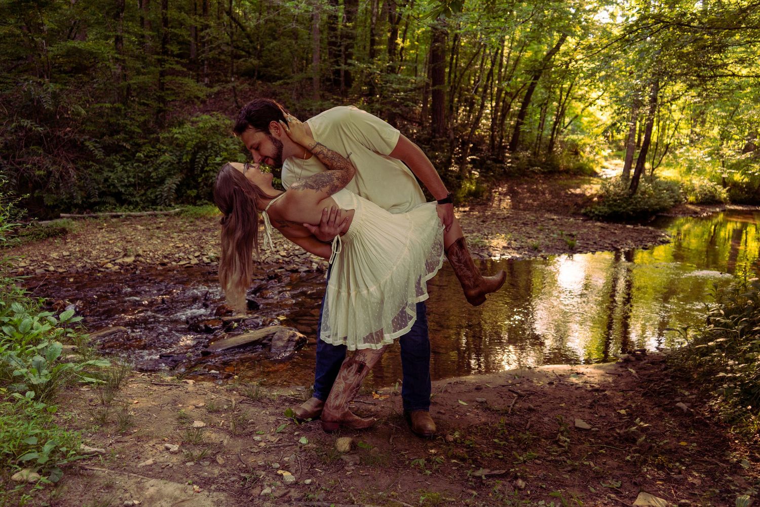 Couple sharing a romantic dip kiss beside a reflective woodland pond surrounded by trees.